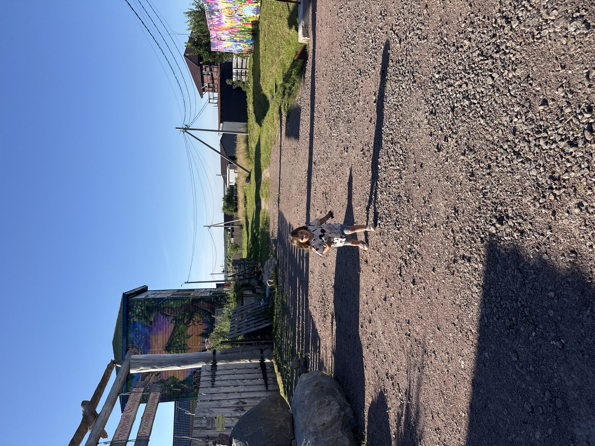 A young girl with dark hair, wearing a light-colored t-shirt with a graphic print and grey shorts, runs along a gravel path towards the viewer. The path is lined with sparse grass and gravel on either side. To the left of the path, there is a large grey boulder and a wooden fence with weathered planks. Behind the fence, a building is visible with a colorful mural depicting a dragon and trees. A wooden sign with Cyrillic lettering hangs from a beam above the fence. To the right of the path, there is a grassy area with more colorful murals on a low structure. In the background, there are utility poles with wires, several houses, and a clear blue sky. The scene appears to be in a rural or semi-rural setting.
