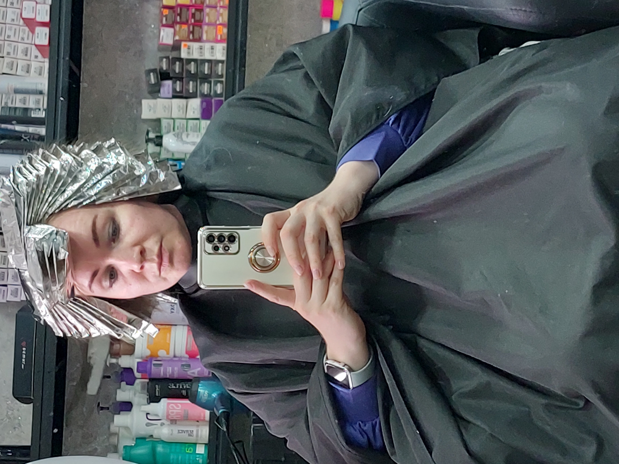 An indoor, eye-level shot captures a person, likely female, undergoing a hair coloring or highlighting treatment in a salon in Chaikovsky, Russia. Her hair is extensively covered with silver foils, fanned out across the top and sides of her head. She is draped in a black salon cape, worn over a blue long-sleeved shirt, which is visible on her left arm.
The person holds a light-colored smartphone, featuring multiple rear cameras and a golden ring holder, in her right hand, looking directly at the camera with a neutral expression. A silver smartwatch with a light-colored band adorns her left wrist.
The background, which appears to be a reflection in a mirror, showcases shelves stocked with numerous hair salon products. These include various boxes of hair dye, some displaying numbers like "10/8" or "8/8" and the brand "L'ANZA HEALING HAIR COLOR". Bottles of different sizes and colors (white, purple, pink, orange, green) are also present, with text like "1882" on a pink bottle and "L'ANZA" partially visible on a green one. A blue hairdryer is partly visible on the right, and a black rectangular object, possibly an organizer with "DEMERD" or similar text, is on the left. The scene clearly depicts a professional hair salon environment.