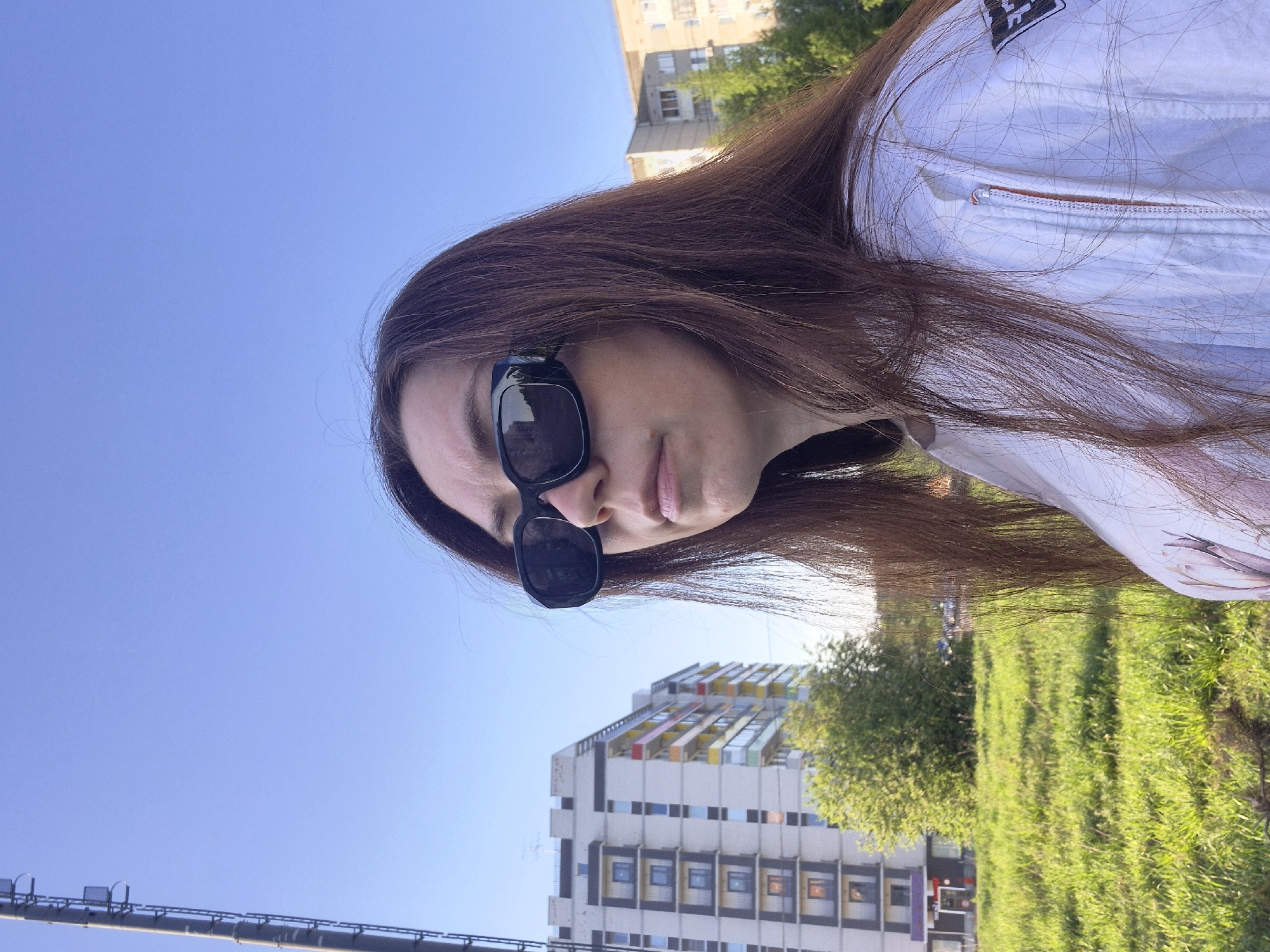A woman wearing sunglasses is in the foreground, with a building and green space in the background. The woman has long, brown hair and is wearing a white jacket. The building behind her is tall and appears to be an apartment building with colorful accents. Green grass and trees occupy the space between the woman and the building. The sky above is clear and blue.