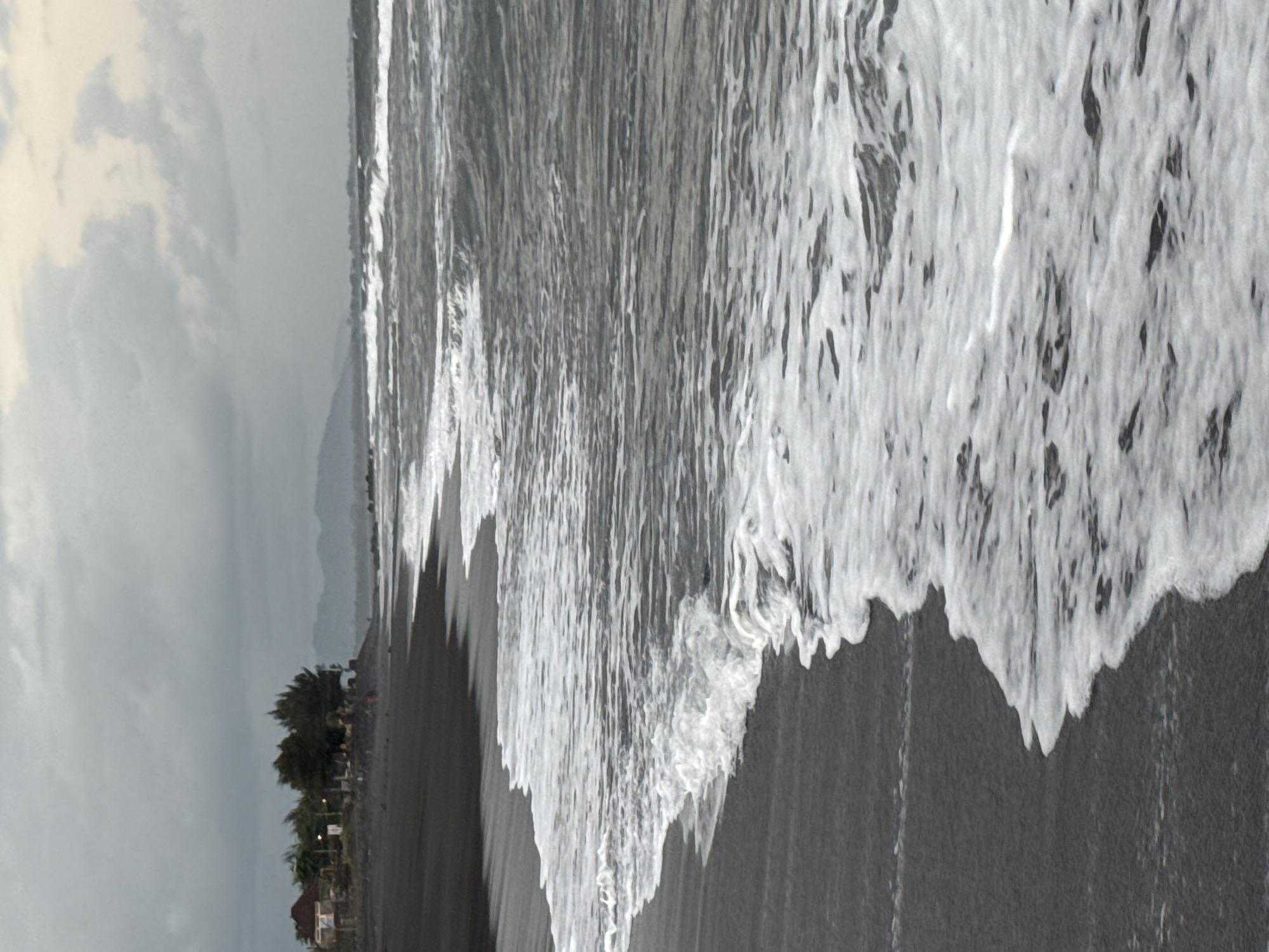 A photograph captures a beach scene on a cloudy day, likely in Blahbatuh, Indonesia. The foreground is dominated by dark sand, with a foamy white wave gently lapping at the shore. In the middle ground, the ocean stretches out, with more waves cresting and breaking. A distant shoreline is visible, featuring a few buildings nestled amongst trees, and further in the background, faint mountains rise against the overcast sky. The overall mood is serene and slightly muted due to the cloudy weather.