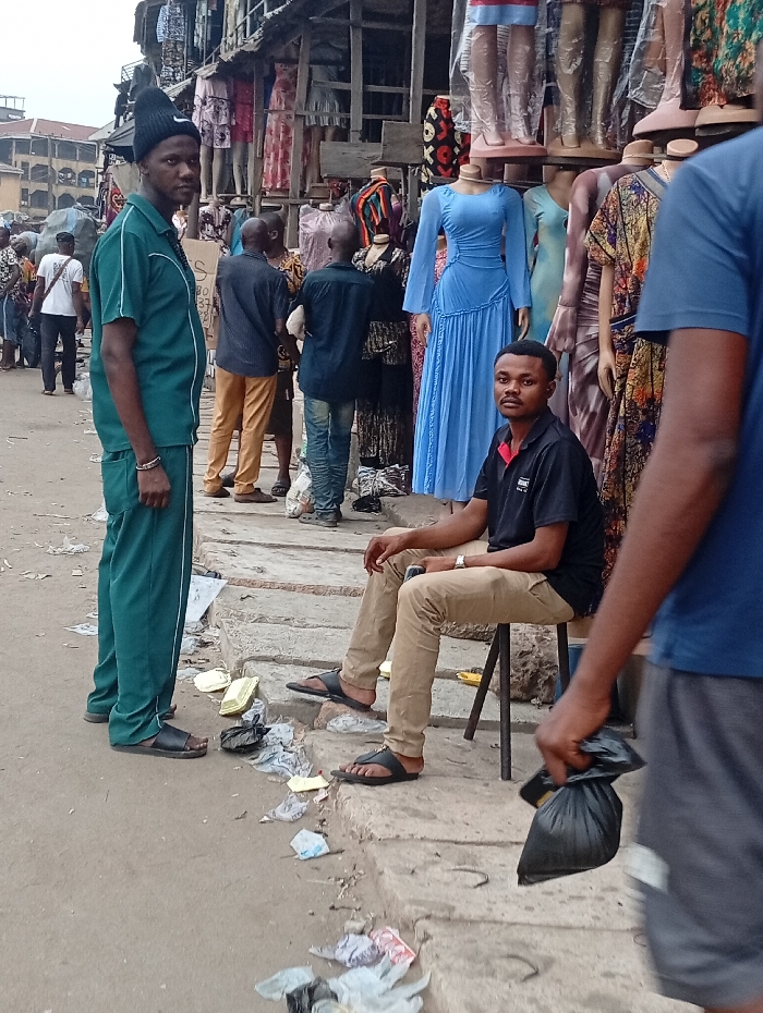 A bustling outdoor marketplace scene depicts two men in the foreground. One stands in a green tracksuit and black beanie, gazing at the camera. Another man sits on a stool, wearing a black shirt and khaki pants. A third man passes by on the right, carrying a black plastic bag. Mannequins dressed in colorful garments line the backdrop of the market, with people browsing and walking in the distance. Litter and debris are scattered along the curb.