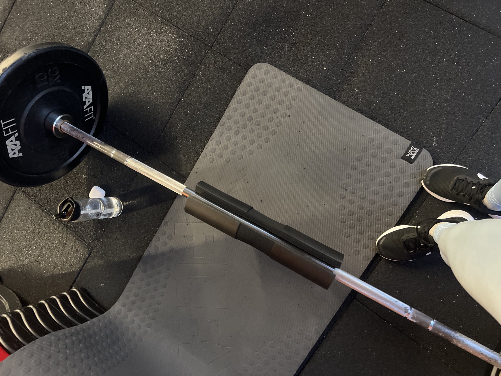 The image shows a top-down view of a gym floor in Plovdiv, Bulgaria. The floor is made of black rubber tiles. A grey exercise mat is placed on the floor. A barbell with a 10 kg weight plate on one end rests on the mat. The barbell has padding in the center. A small, clear water bottle sits next to the barbell. A pair of black athletic shoes are visible near the edge of the mat, indicating a person is present but only partially visible.