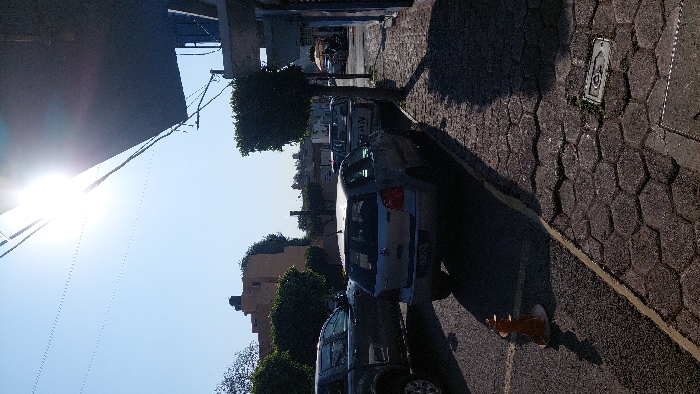 This image depicts a street scene in Tlaxcala City, Mexico, under direct sunlight from the upper right, causing lens flare and strong shadows. The street is asphalt with a yellow painted line marking the edge of a parking area. An orange, cone-shaped plastic traffic delineator with a white base rests on the asphalt in the lower-left foreground. Several vehicles are parked: a dark grey pickup truck is partially visible on the far left, followed by a silver Volkswagen Polo hatchback (MK4 generation) facing away from the camera. Behind the Polo, a white Nissan pickup truck is parked, displaying "NISSAN" on its tailgate. The sidewalk to the right consists of dark hexagonal pavers, with a metal utility cover embedded in the lower right. A small, trimmed tree stands on the sidewalk between the parked cars and a dark blue building facade, partially visible under a dark awning that extends into the upper right. In the background, other buildings are visible, including a light brown structure with a rooftop water tank on the left. The sky is clear and light blue. No individuals are clearly visible.