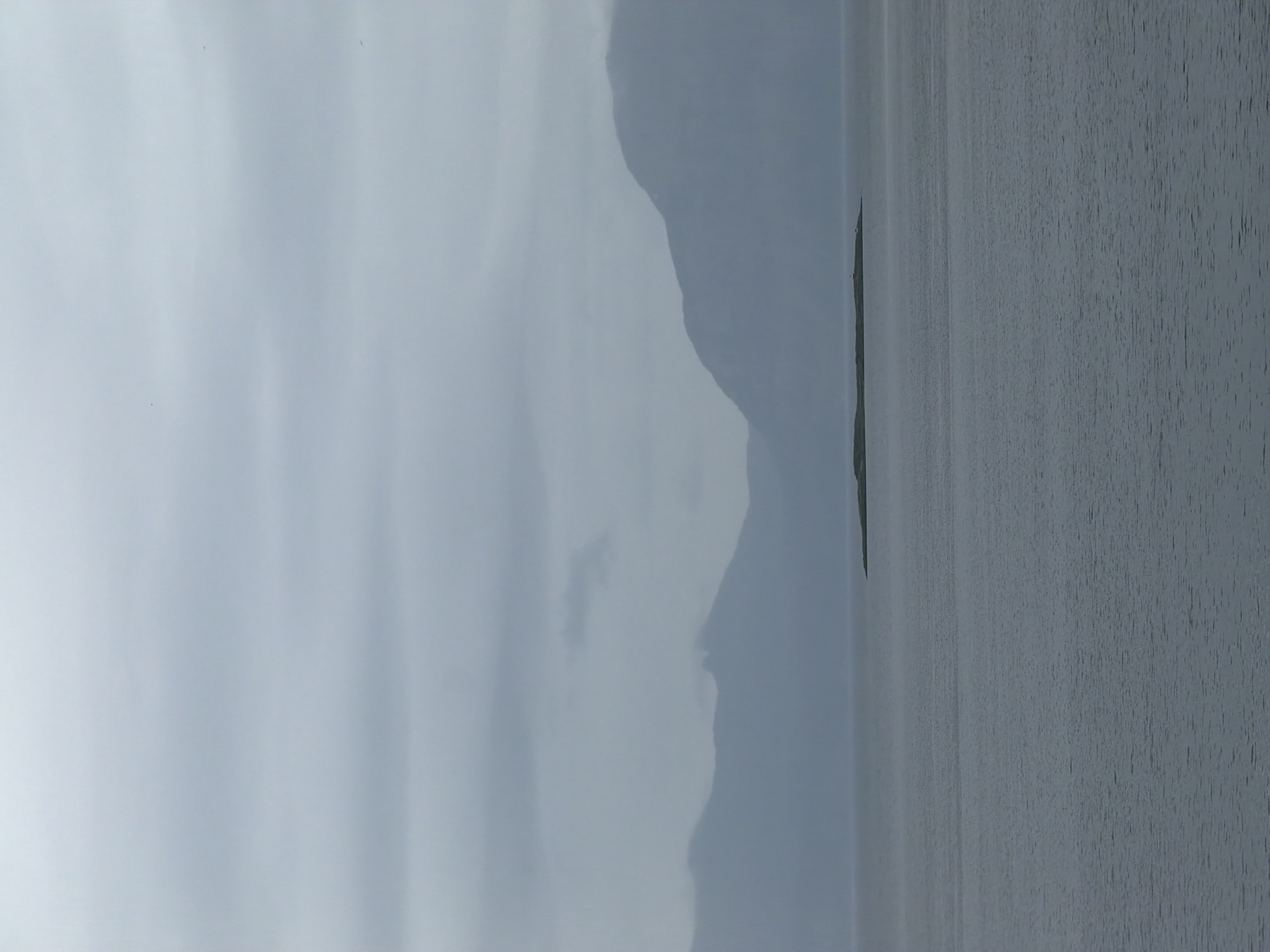 The image depicts a broad view of a calm body of water, likely a fjord or sea, with a low-lying island situated in the middle distance. The water is a uniform grey, with subtle ripples indicating minimal movement. Behind the water, a series of muted, hazy mountain ranges dominate the horizon, their peaks softly defined against the overcast sky. The sky itself is a uniform, pale grey, suggesting a cloudy or foggy day. The overall impression is one of a tranquil, perhaps somewhat melancholic, natural landscape. The location is likely the waters surrounding Harstad, Norway, given the characteristic mountainous coastal scenery. There are no people or specific structures visible on the island or in the surrounding water, focusing the scene solely on the natural elements.