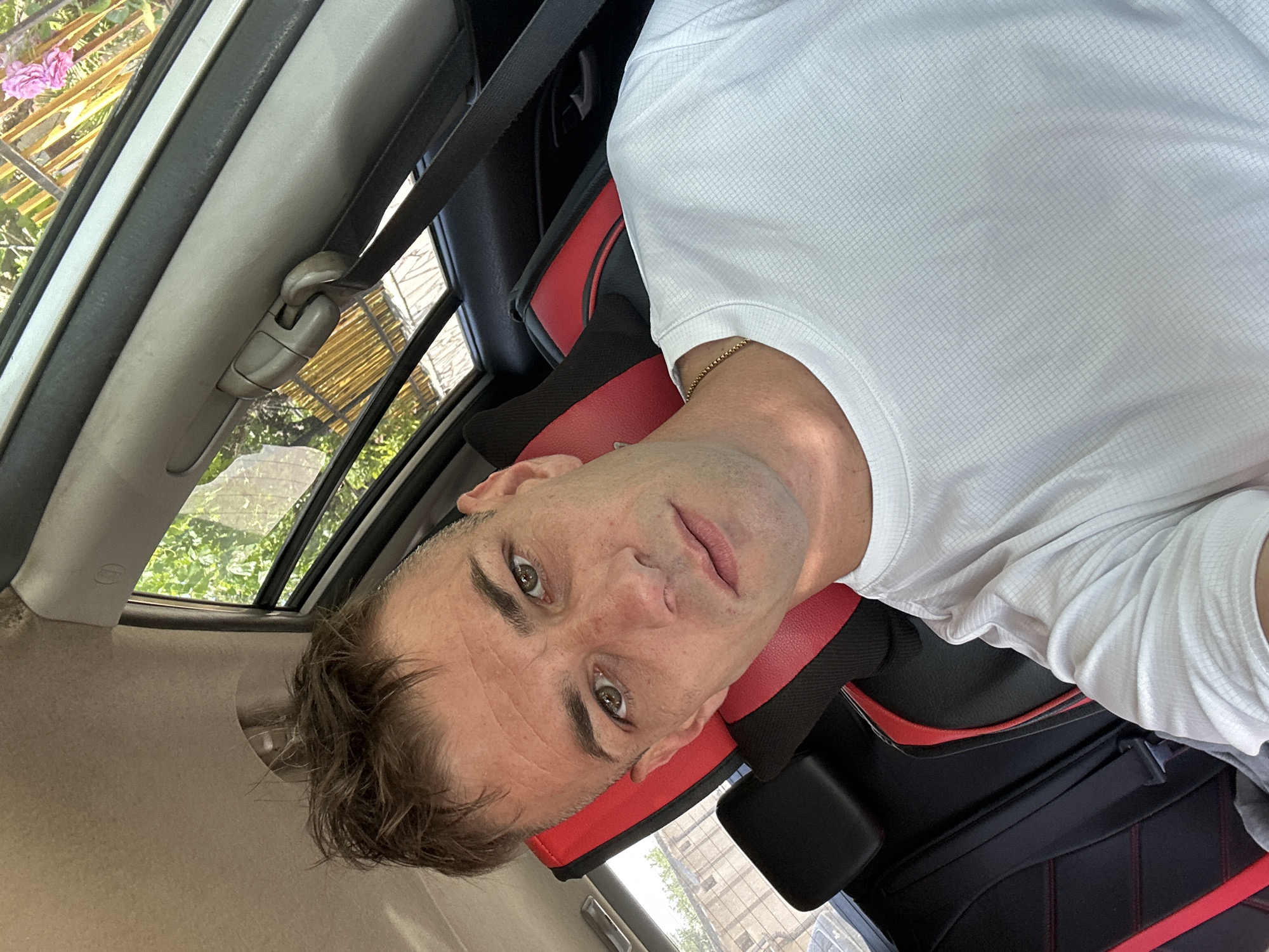 A man is taking a selfie inside a car. The man has short, messy brown hair, green eyes, and is wearing a white t-shirt and a gold chain. He is looking directly at the camera with a neutral expression. The interior of the car is visible, including the black and red seats and a seatbelt. Outside the car window, trees and a golden fence are visible. The scene appears to be in Cherven Breg, Bulgaria, as indicated by the prompt, though there are no explicit visual cues to confirm this location.