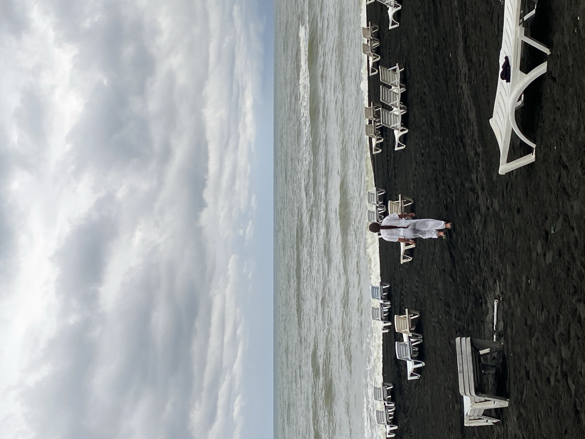 The image depicts a cloudy day at Ureki, Georgia's black sand beach. A person in white clothing walks away from the camera towards the sea. The beach is populated with numerous empty white lounge chairs, some of which are stacked. The dark sand contrasts with the white furniture and the grey, choppy water of the sea. The sky is overcast with heavy, layered clouds.
