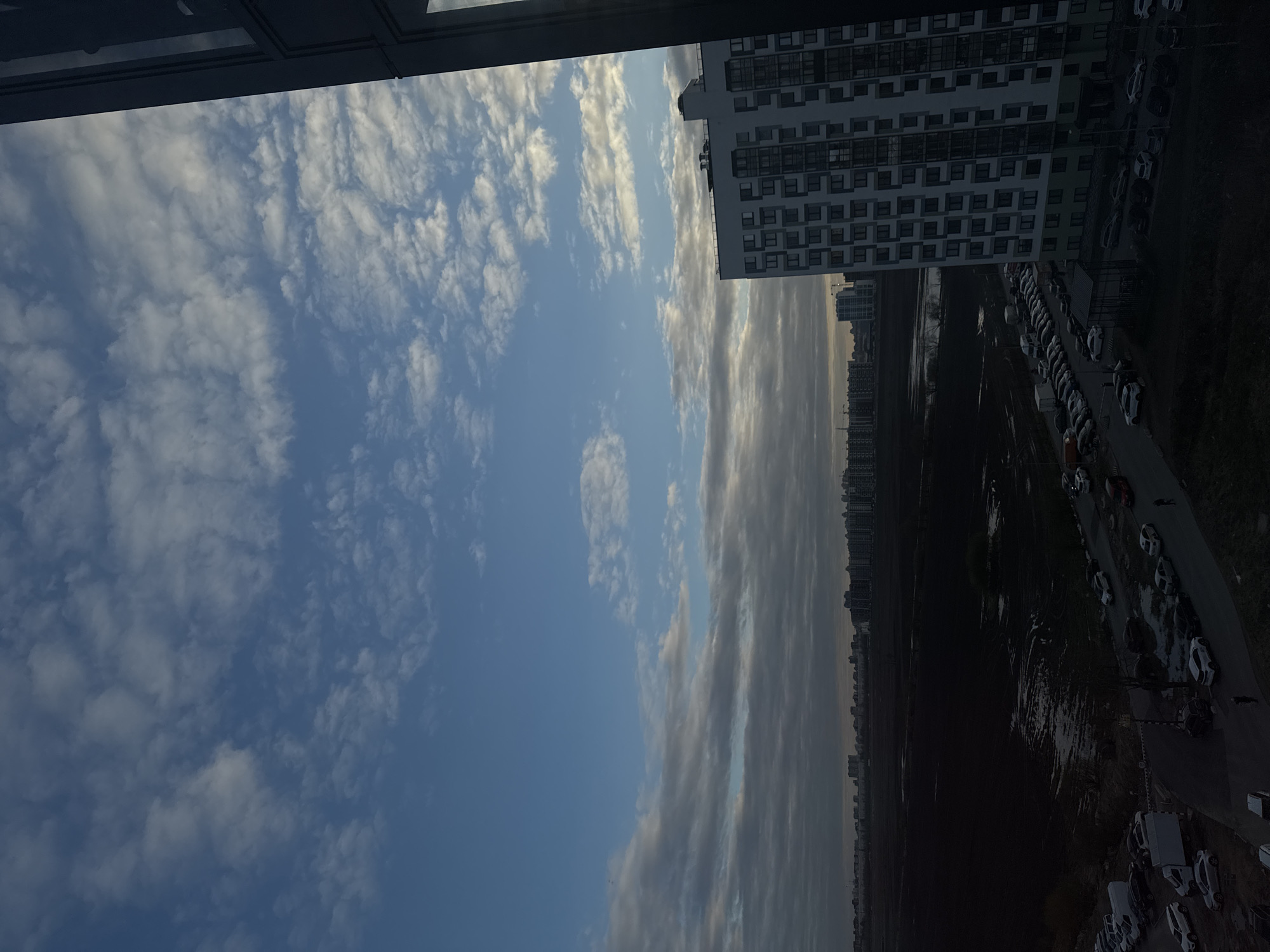 The image, taken from a high vantage point, presents an urban or suburban landscape in Saint Petersburg, Russia, under a partly cloudy sky. A dark, possibly metallic, frame or structure is visible at the very top, indicating the photograph was taken from an enclosed space like a window or balcony.
The upper and central parts of the image are dominated by the sky, displaying a clear blue interspersed with numerous white and light-grey cumulus and stratocumulus clouds. The clouds show differential illumination, suggesting a low sun angle, characteristic of either early morning or late afternoon.
To the right, a significant portion of a modern, multi-story apartment building is visible, characterized by a light-colored facade and numerous dark, rectangular windows arranged in a grid pattern. Below this building and extending across the lower half of the frame is a dark, expansive area that appears to be a mix of undeveloped land, possibly a muddy or wet field, or a body of water reflecting the dim light. Adjacent to this area, a long row of cars is parked, predominantly white or light-colored, indicating a parking lot or a street.
In the far distance, along the horizon, a faint silhouette of a city skyline with additional buildings can be seen, further emphasizing the urban setting. The overall lighting casts long shadows and creates a subdued atmosphere, particularly over the ground and distant structures. No specific human activity is depicted, and no text is visible.