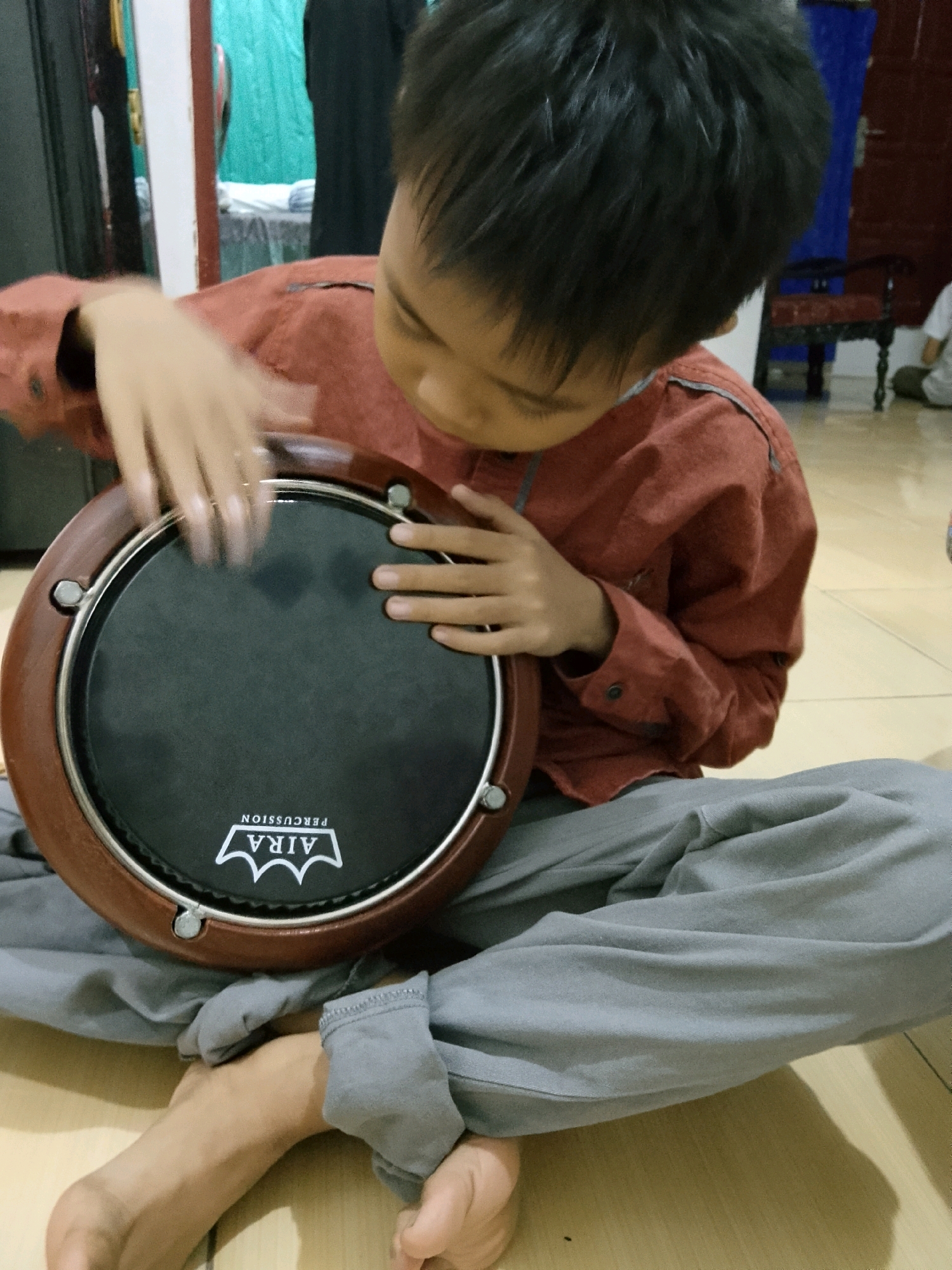 An Asian boy, appearing to be around 7-9 years old, sits cross-legged on a wooden floor, playing a frame drum. He is wearing a rust-colored, long-sleeved shirt and grey pants. His hands are blurred, indicating movement as he strikes the drum. The drum has a brown wooden rim and a black playing surface with a white logo that reads "AIRA PERCUSSION". The background is slightly out of focus, showing a room with a teal curtain, a table with some items on it, and furniture. The lighting appears to be natural, coming from an unseen window or light source. The image is taken from a slightly elevated angle, looking down at the boy and the drum. The scene suggests an indoor setting, possibly a home or a cultural center in Palembang, Indonesia, where such musical instruments are common.
