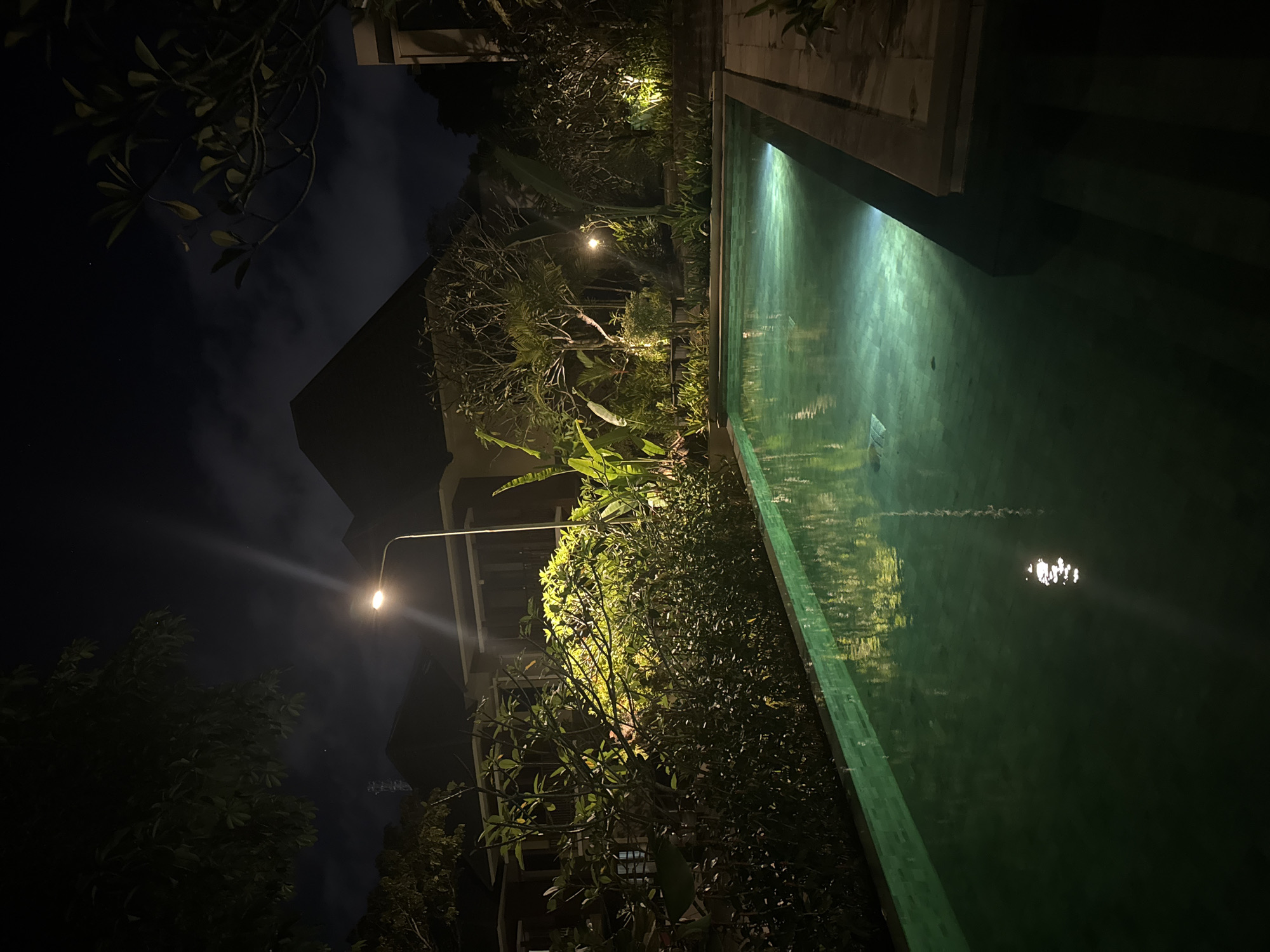 The image shows a swimming pool at night in what appears to be Kuta Selatan, Indonesia. The pool is rectangular and occupies the right side of the frame. The water surface reflects the surrounding light, creating a shimmering effect. The pool's edge is visible, constructed of light-colored material. The left side of the image is dark, with foliage and trees illuminated by scattered lights. The outline of a building is partially visible in the background, adding to the tropical night setting. A street lamp casts a strong glare in the direction of the camera lens.