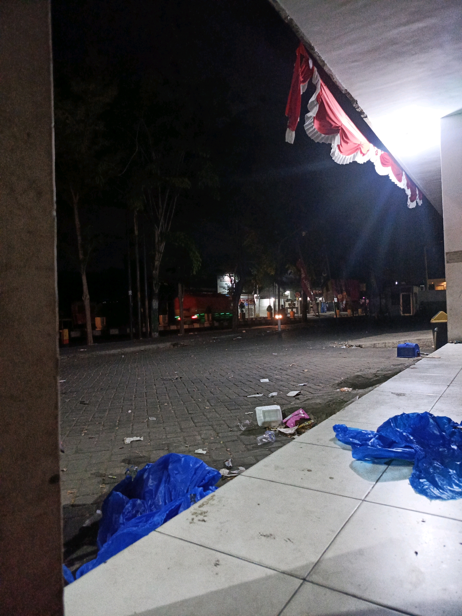 The image depicts a street scene at night in Pamekasan, Indonesia, likely in a commercial or public area. The foreground is dominated by a tiled walkway with several pieces of blue plastic trash bags and some scattered debris. To the left, a textured pillar is visible. Beyond the walkway, a paved street is strewn with various types of litter, including plastic containers and paper waste. Trees line the side of the street, and in the background, there are buildings and several vehicles, including a red truck with its lights on. A red and white decorative banner is draped from an overhang at the top right of the image, illuminated by a bright light source. The overall impression is one of a somewhat neglected urban environment after dark, possibly after an event or at the end of a business day.