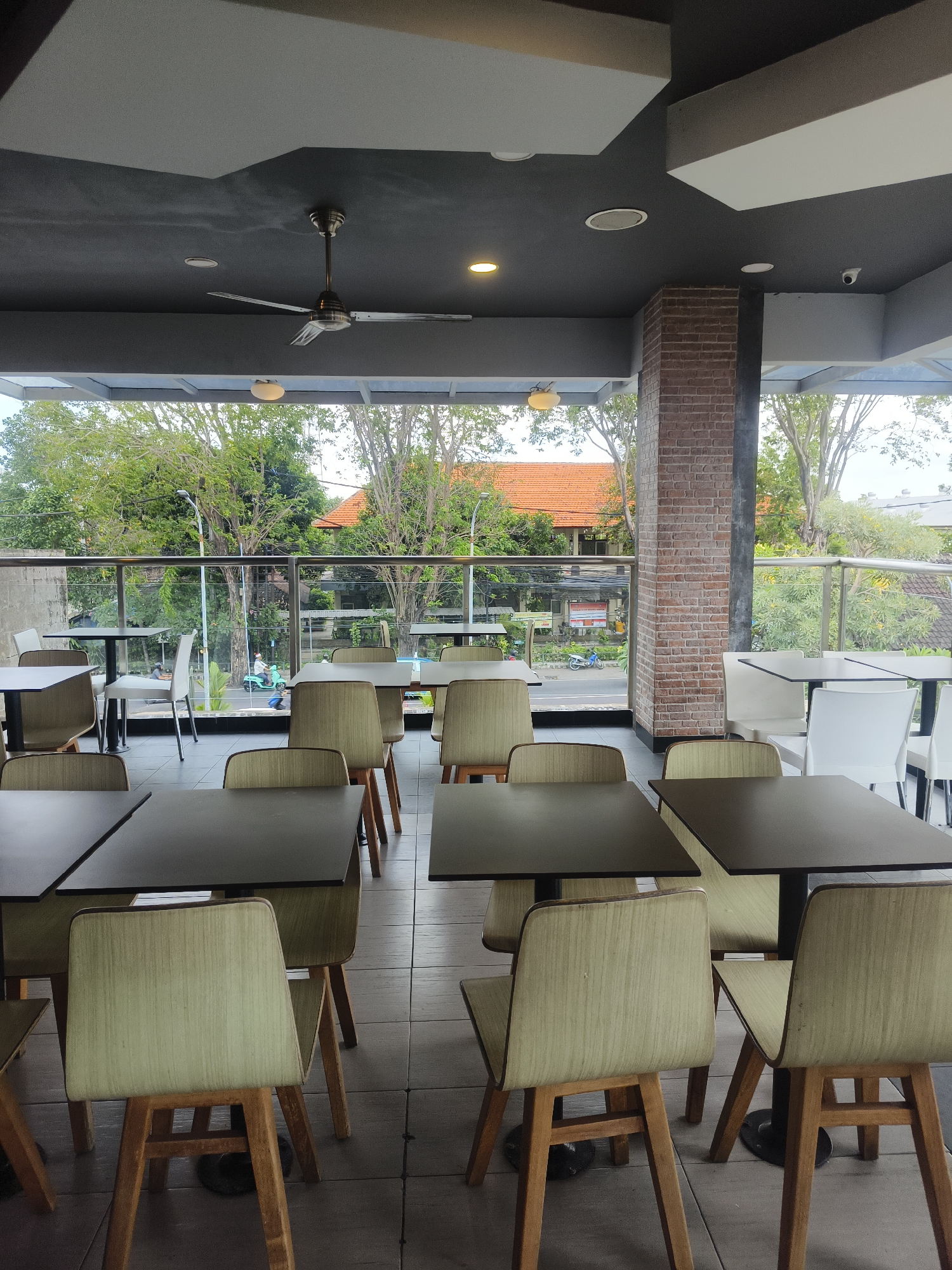 The image depicts an empty outdoor seating area of a cafe or restaurant, likely in Denpasar, Indonesia, given the lush green trees, reddish-tiled roofs, and a scooter with a rider visible through the glass partition. The seating arrangement consists of multiple dark square tables paired with chairs. The chairs are upholstered in a light beige fabric and have wooden frames and legs. The flooring is tiled in a neutral grey. Above the seating area, a dark grey ceiling features a ceiling fan and recessed lighting. A brick pillar is visible on the right side of the frame. The background shows a street scene with more greenery and buildings. The overall impression is of a quiet, possibly late afternoon, atmosphere, with the seating area waiting to be occupied.
