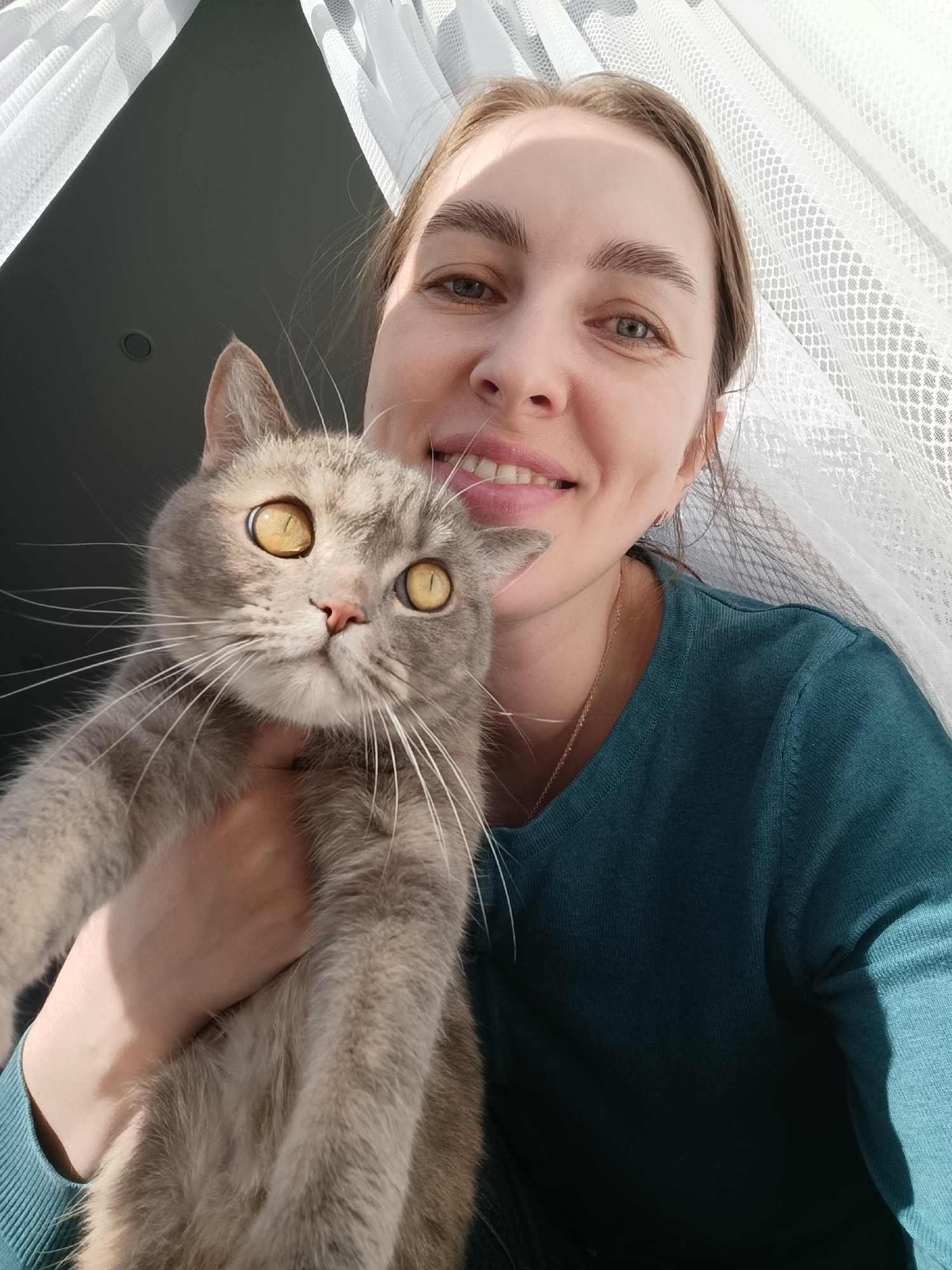A light-skinned woman is holding a grey cat in her arms, both facing the camera. The woman is smiling, showing her teeth. She has light brown hair pulled back and is wearing a dark teal long-sleeved shirt and a gold chain necklace. The cat has yellow eyes and a pink nose. Behind them is a translucent white curtain hanging in front of what appears to be a window. The location is Chaikovsky, Russia.