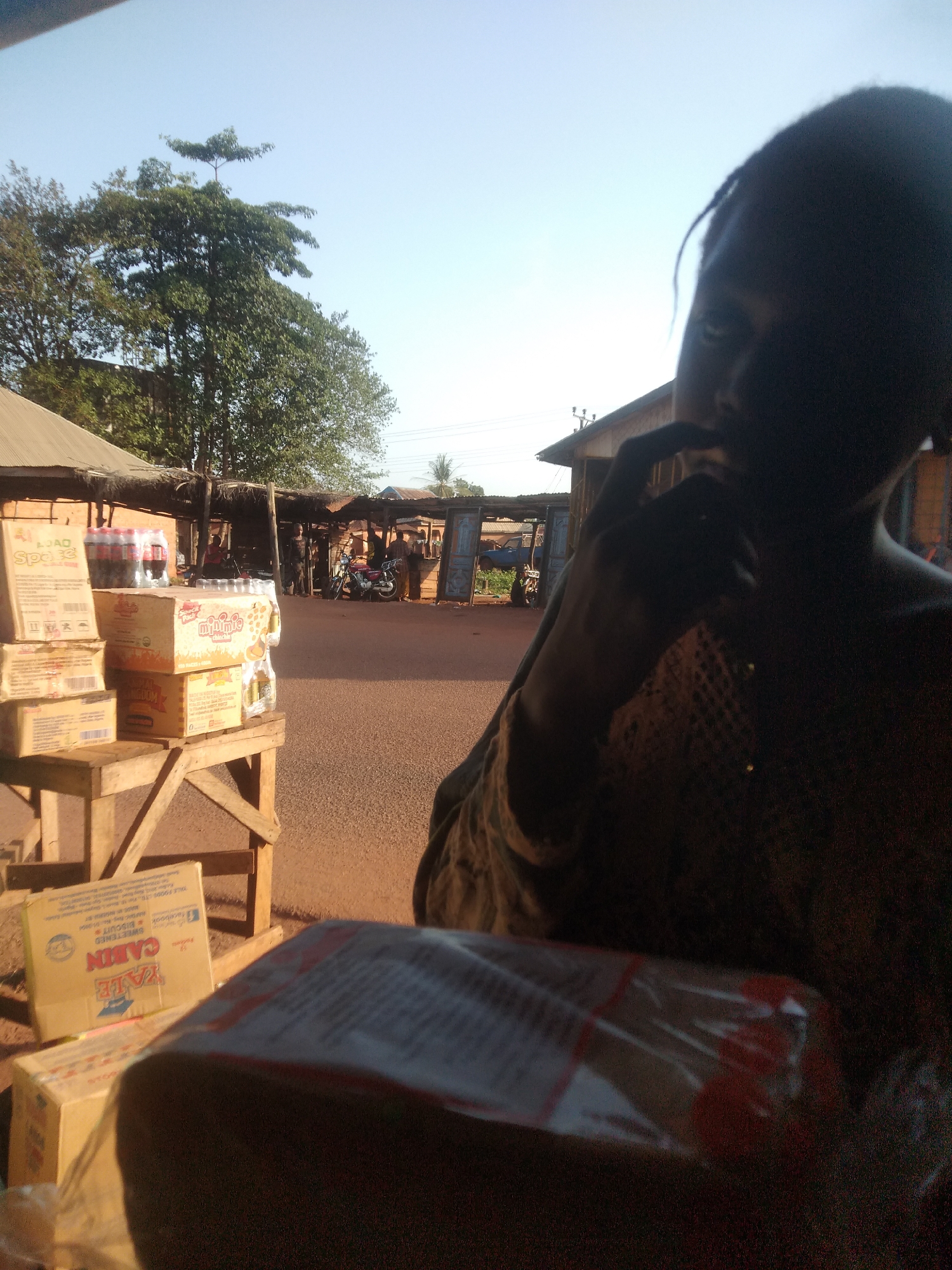 The scene appears to be a market area in Ejule, Nigeria. In the foreground is a dark-skinned person with their hand near their face; their features are obscured by shadow. A cellophane-wrapped item is in the very front. Slightly further back and to the left, a wooden stand displays several boxes of food items including "Space" and "Minimie Chin Chin". Soft drinks are also visible. Behind this stand is a street scene with people, motorcycles, and corrugated metal buildings indicative of a trading area. The ground is reddish-brown. Trees line the street, and the sky is clear blue.