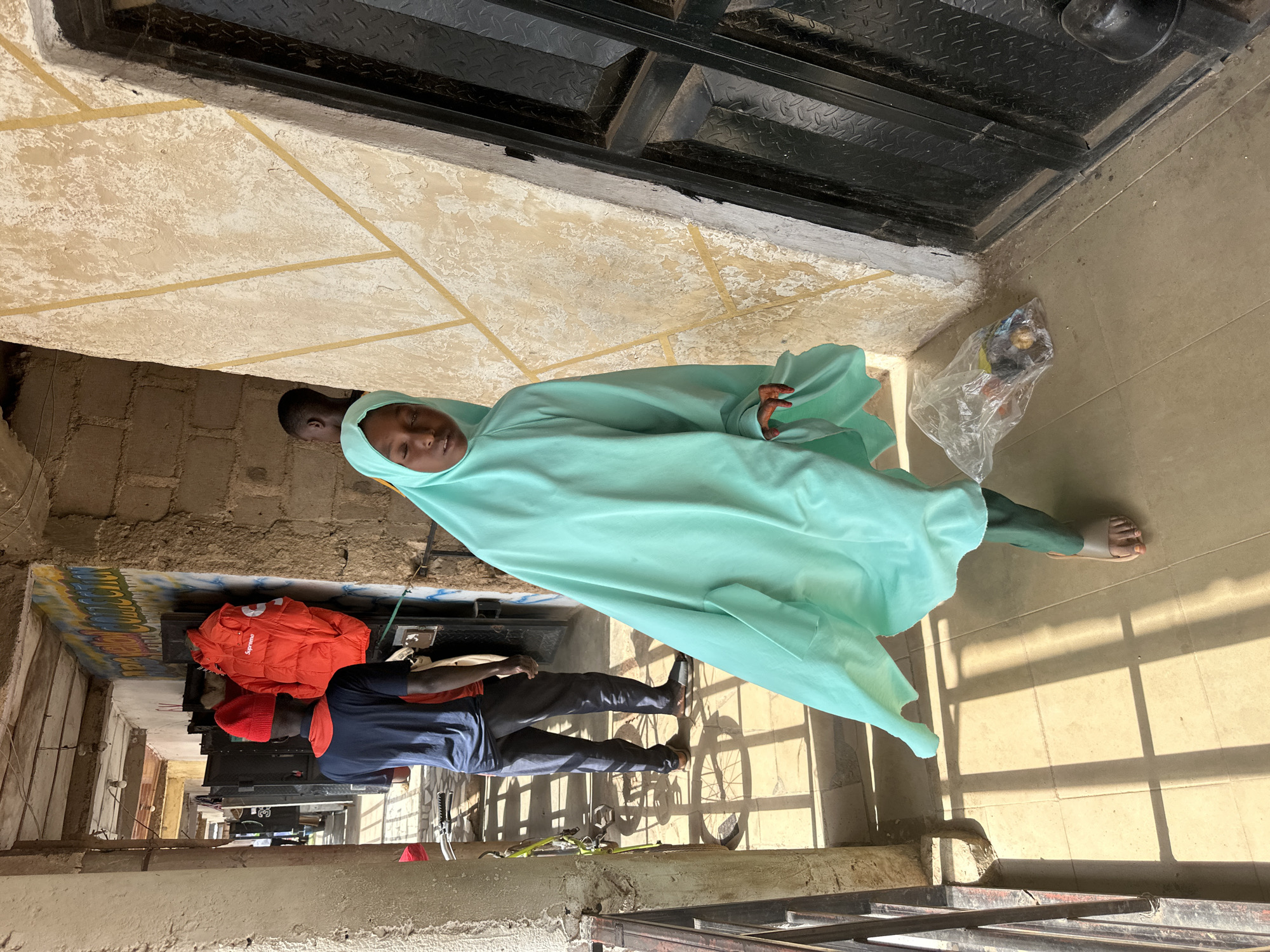 In Bukuru, Nigeria, a young girl wearing a light green hijab and a flowing gown walks down a covered walkway. She is carrying a transparent plastic bag filled with items. To her left, a man in a dark blue t-shirt and gray pants with a red shirt underneath is standing near a bright orange jacket hanging on a wall adorned with graffiti. Another person is partially visible behind the girl. The walkway has a tiled floor with sunlight casting shadows from a railing on the left. The walls are made of concrete blocks and stucco, with a black metal door on the right.