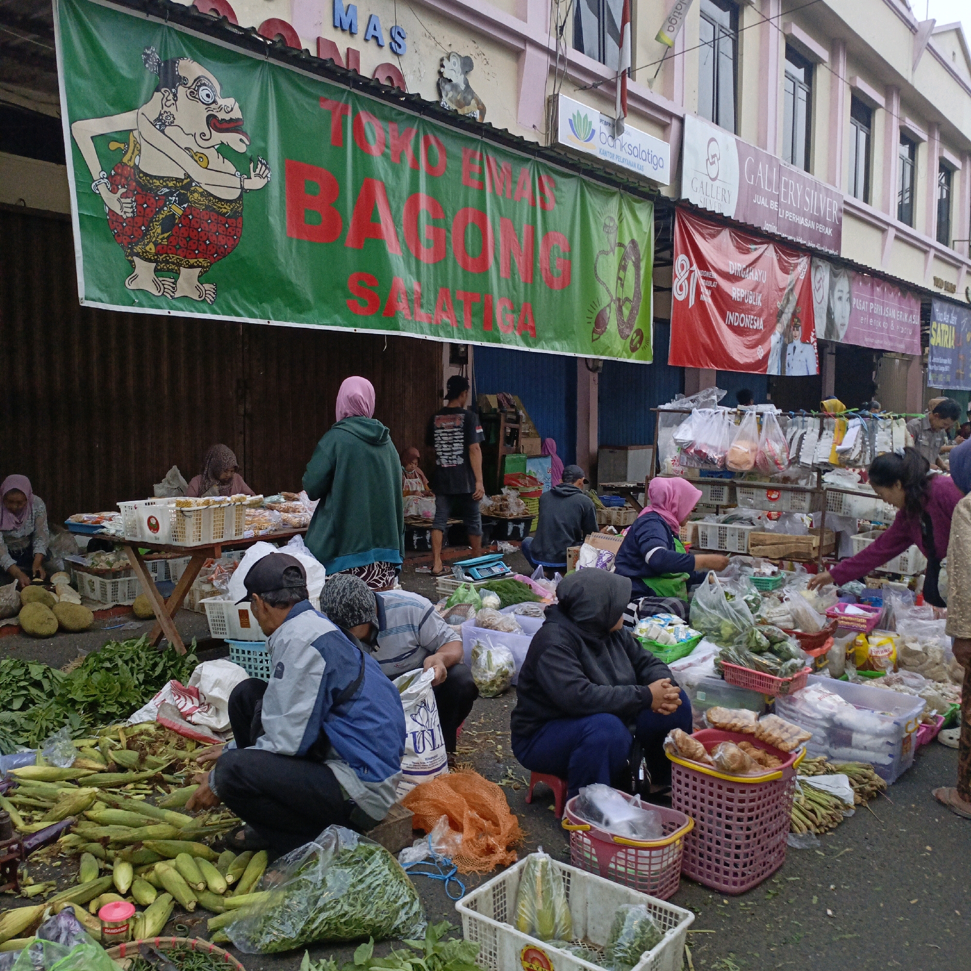 The image depicts an outdoor market scene in Salatiga, Indonesia, likely in front of a row of shops. A large green banner with red lettering, featuring a cartoonish figure and the text "TOKO EMAS BAGONG SALATIGA", hangs prominently overhead, indicating the name and location of a business. Various vendors and customers are engaged in commercial activity. In the foreground and midground, individuals are seen behind tables and on the ground, surrounded by piles of fresh produce, including corn, eggplants, and leafy green vegetables, displayed in baskets and plastic bags. Some people are sitting or squatting, interacting with the goods or with each other. The background shows the facades of buildings with windows and some signage, suggesting a commercial street setting. The overall atmosphere is that of a bustling local market.