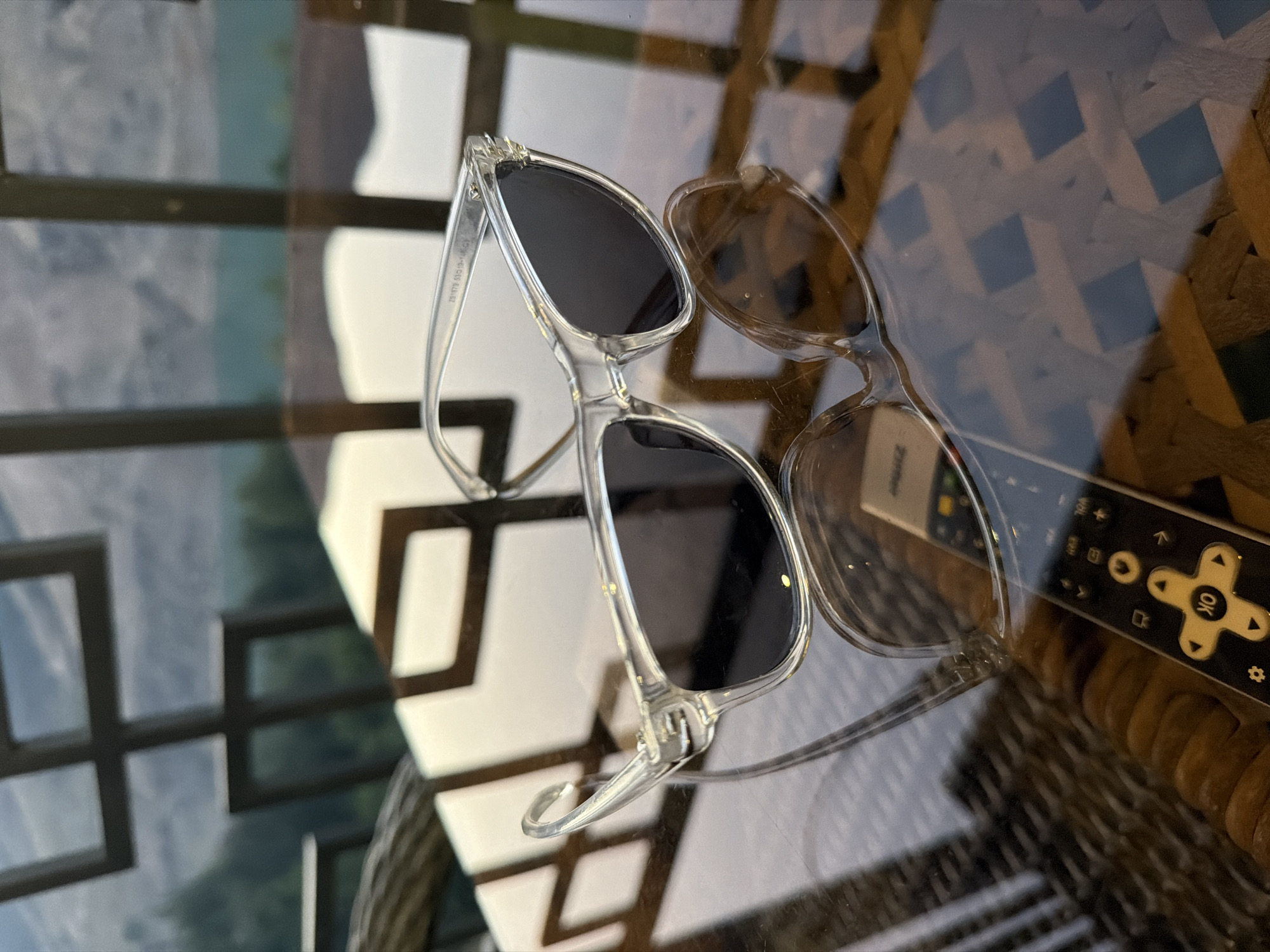 A pair of clear-framed sunglasses with dark lenses rests on a reflective glass tabletop. The sunglasses are positioned at an angle, with their reflection visible below. A black remote control with yellow buttons is partially visible beneath the sunglasses, also reflected on the table. The tabletop's surface has a woven pattern, and beyond it, blurred greenery and a body of water are visible, suggesting an outdoor setting. The background features a dark, geometric structure. The image appears to be taken at an eye-level perspective. There are no people or animals present in the scene. The location is not specified in the image, and therefore cannot be confirmed to be in the city of Nanay, Uzbekistan.