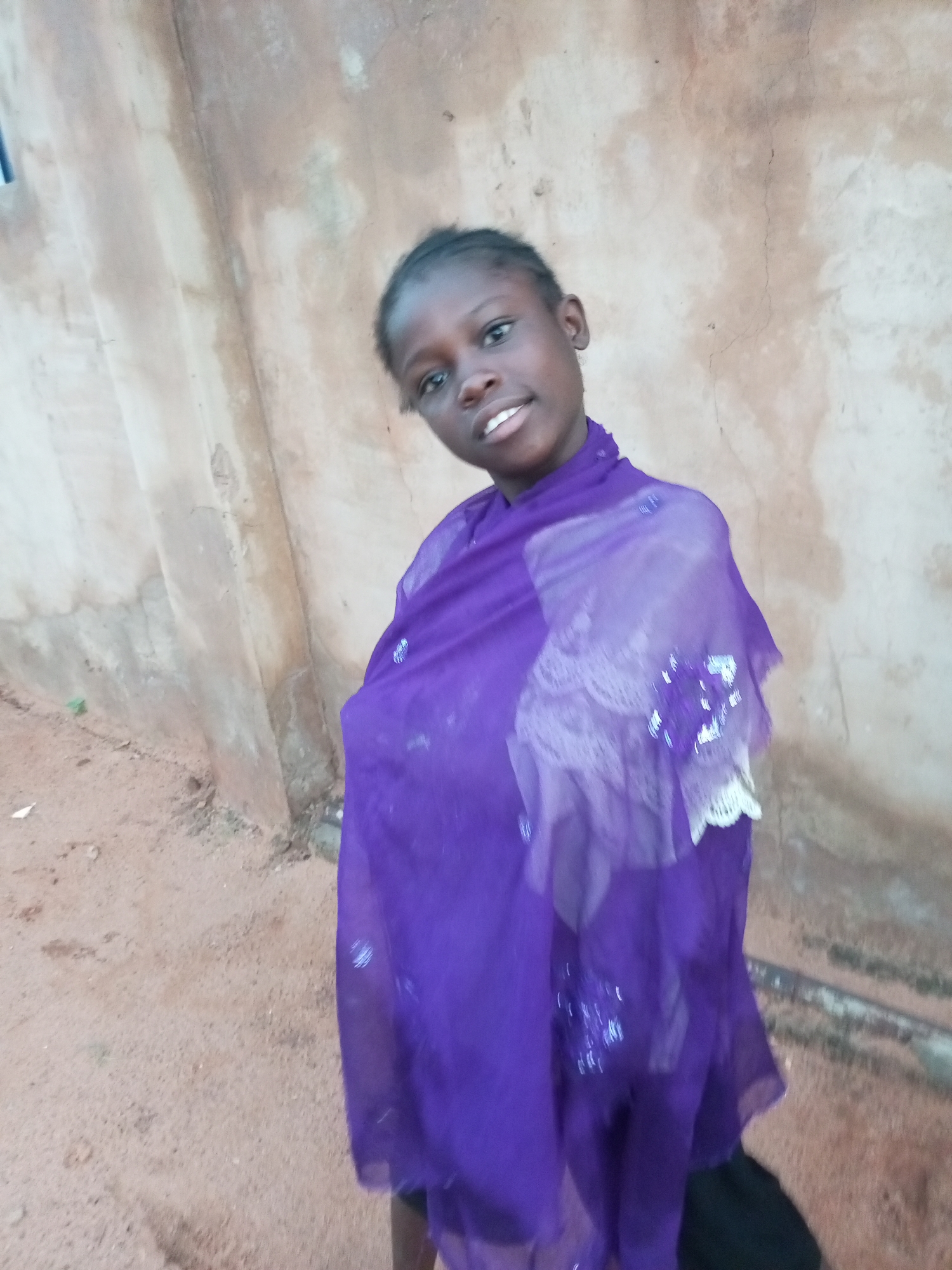 A young girl stands in front of a textured, light-brown wall. She is wearing a vibrant purple sheer scarf draped over her shoulders, which is adorned with white lace and some sparkling embellishments. Her expression is friendly, with a slight smile. The ground in front of the wall appears to be sandy or dirt. The scene is outdoors.