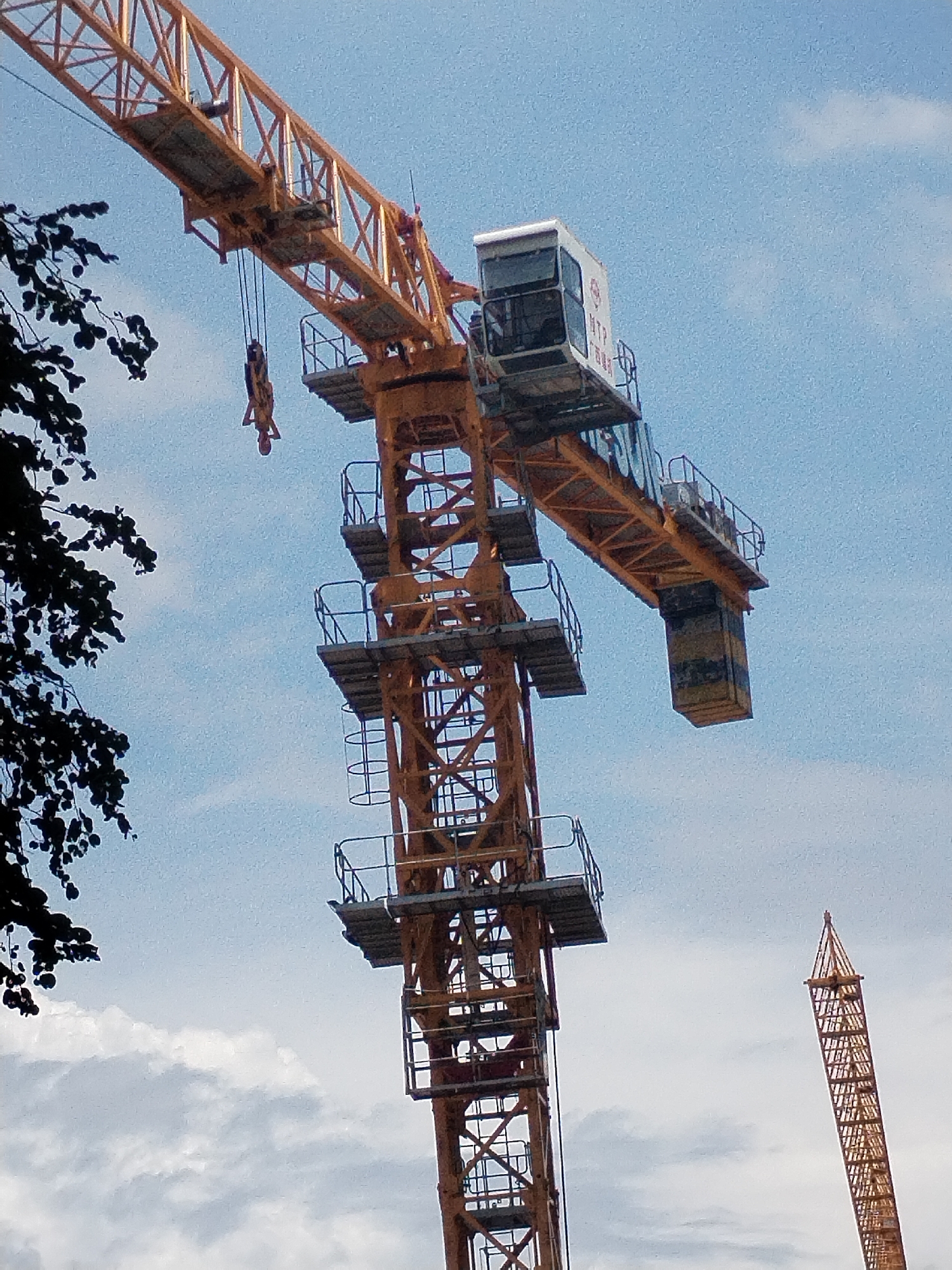 This is a low-angle, medium shot of a construction crane against a bright blue sky with scattered clouds. The primary crane is large and orange, with a visible operator's cab at the top of its vertical tower. A horizontal jib extends from the tower, with a hook and cable hanging down, carrying a suspended object that resembles a person's torso. The construction site appears to be in Nha Trang Ward, Vietnam, indicated by the presence of another, taller, yellow construction crane in the background on the right. Dark green foliage from a tree is visible in the upper left corner, partially obscuring the sky and the crane. The overall scene suggests ongoing construction activity in an urban or developing area.