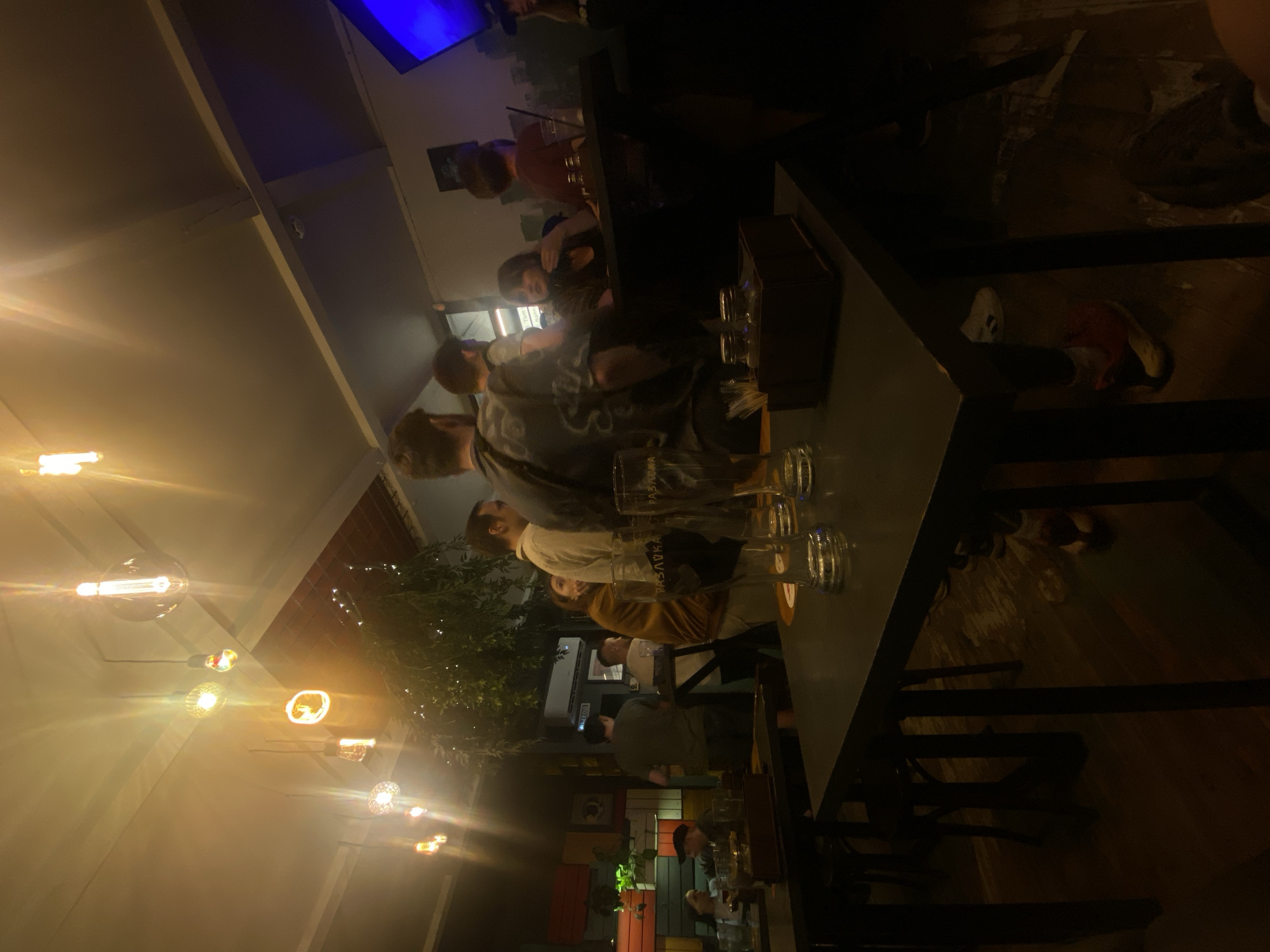 The image captures a dimly lit bar scene, potentially in Заволжье, Russia, featuring a group of people gathered around a high-top table. Several clear glasses, possibly beer glasses, are visible on the table. The people appear to be engaging in conversation, and some are standing while others are seated. String lights are strung overhead, adding to the ambiance. The bar is furnished with dark-colored tables and chairs. The background reveals a hint of decorative foliage and other patrons in the establishment.