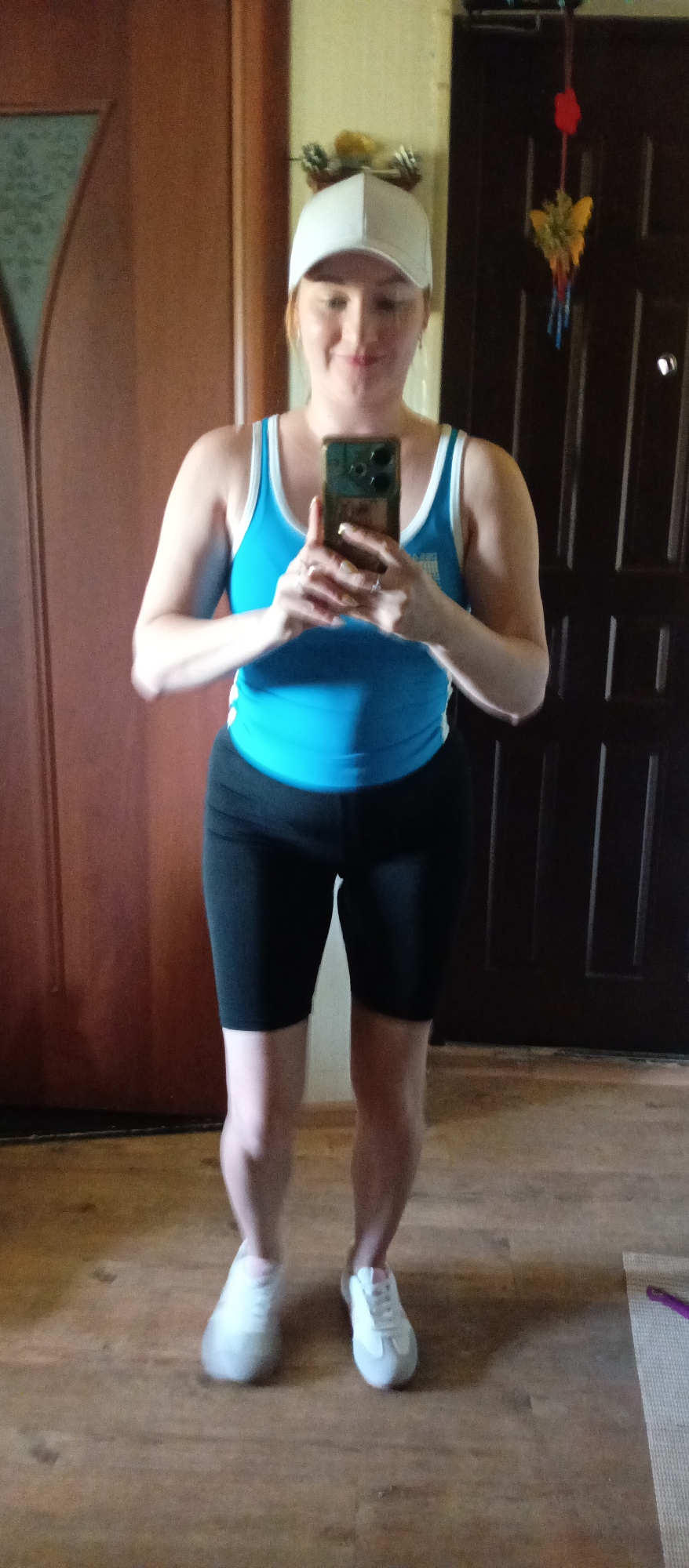 A fair-skinned woman is standing indoors, presumably in a residential setting of an unknown city, as indicated by the lack of recognizable landmarks. She is taking a selfie with a cell phone, the phone held in both hands. The woman is wearing a white baseball cap, a blue tank top with white straps and black athletic shorts. She also has on white sneakers with a grey stripe near the toe. The floor beneath her is wood grain. Behind her are two dark brown doors, one appearing to be a standard interior door and the other possibly a front door with a decorative hanging item.