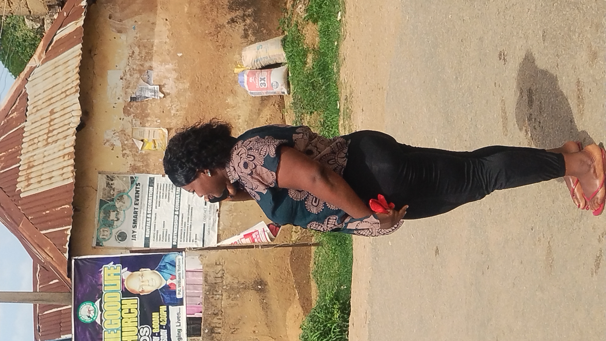 A woman wearing black pants and a patterned shirt is standing on a concrete surface in Kabong, Nigeria. She is wearing red flip-flops and appears to be talking on a cell phone.
Behind the woman is a dilapidated building with a corrugated metal roof. There are several posters affixed to the wall of the building, advertising events and services. One poster advertises "Jay Smart Events," specializing in rentals and decorations. Another poster promotes "The Good Life Church," with a picture of Pastor Williams Tsokwa, inviting people to a service on Sunday at 8:00 AM and Sunday at 4:30 PM.
Next to the building is a patch of green grass. There are two bags of cement leaning against the wall.