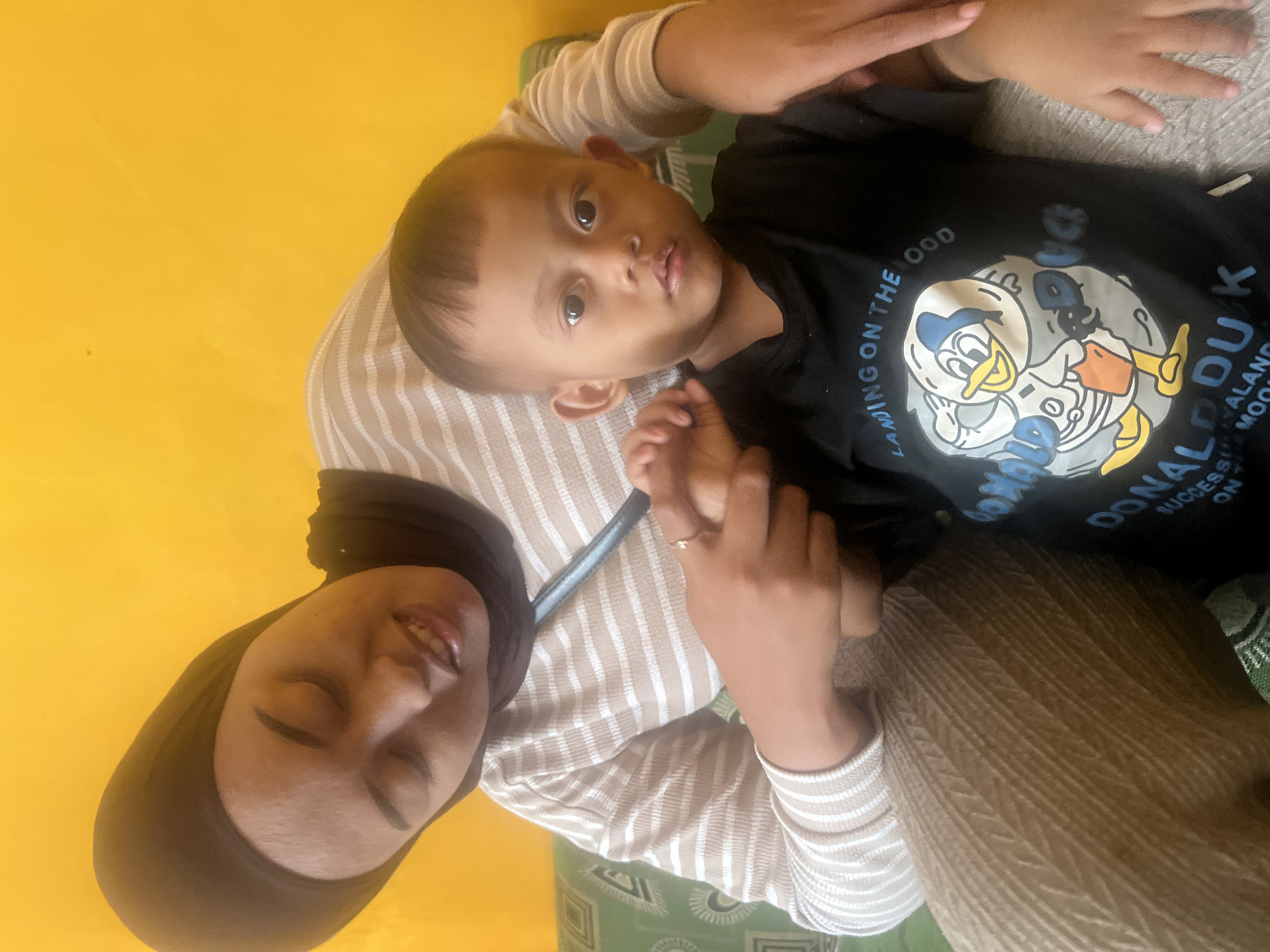 A medium shot shows a woman wearing a hijab and a striped shirt embracing a young boy. The woman's head is tilted down, and she is looking at the boy. The boy is looking forward with wide eyes. The background is a bright yellow wall. The image appears to have been taken indoors. The scene does not provide any specific details about the location in Kecamatan Pati, Indonesia.