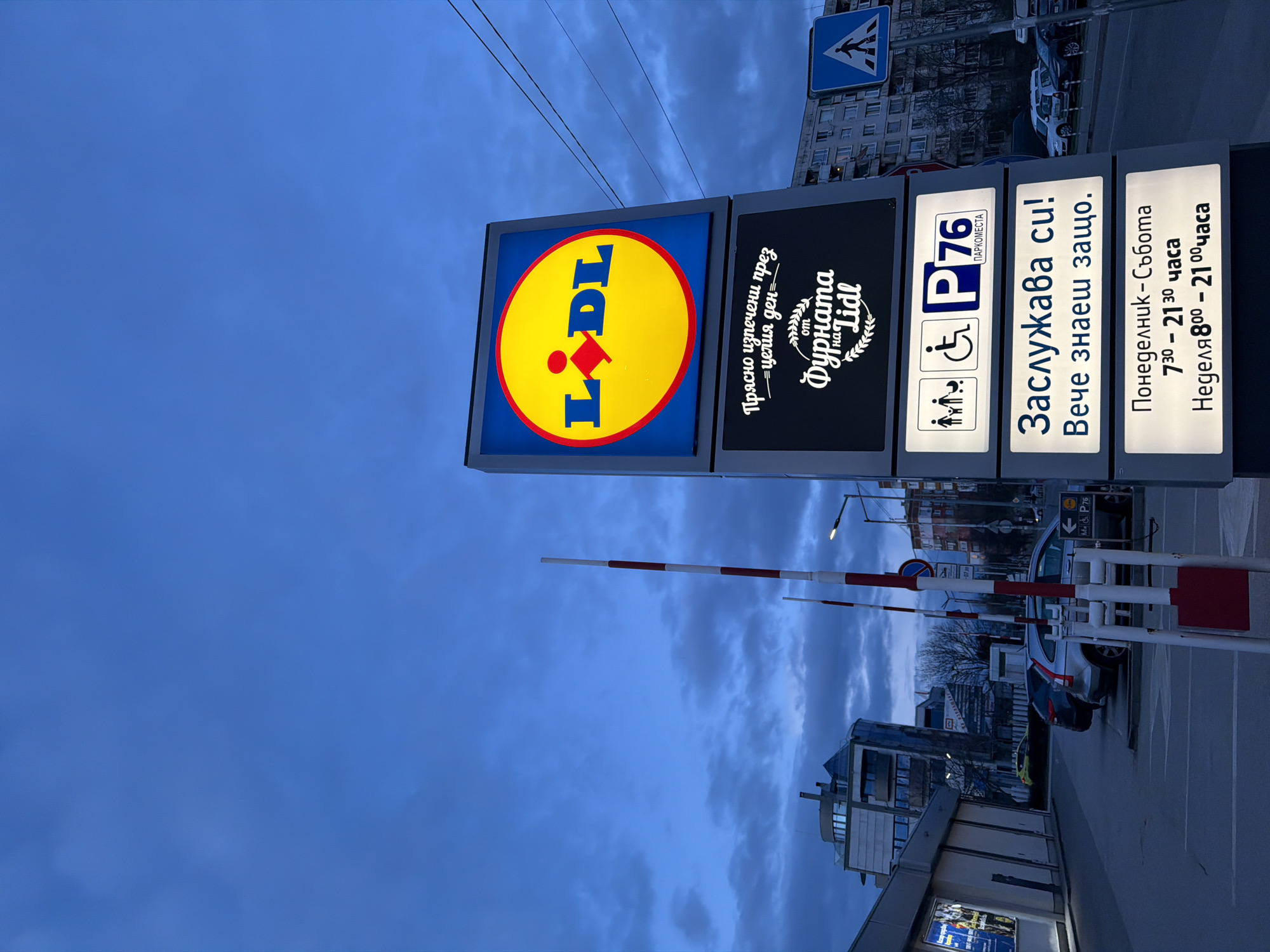 Here is a description of the image:
The image shows a tall, vertical sign for a Lidl supermarket in Sofia, Bulgaria. The sign is made up of several sections. The first and largest section features the Lidl logo: a yellow disc with the word "LIDL" in blue letters and a red oval above the "I." To the right, a black sign reads "Прясно изпечени през = целия ден" and "Фурната на Lidl." The next sign is white and displays symbols for parking, families, and accessibility, along with the text "P76 ПАРКОМЕСТА." The next sign is white with the phrase "Заслужава си! Вече знаеш защо." The final sign lists the store's hours: "Понеделник - Събота 730 - 21 30 часа" and "Неделя 800 – 2100 часа."
In the background, the sky is a deep blue with clouds, suggesting dusk or dawn. A crosswalk sign is also visible. Below the supermarket signs, there is a parking gate barrier with red and white stripes. Cars are parked on the right. The building across the street and the street lamps suggest a business environment.