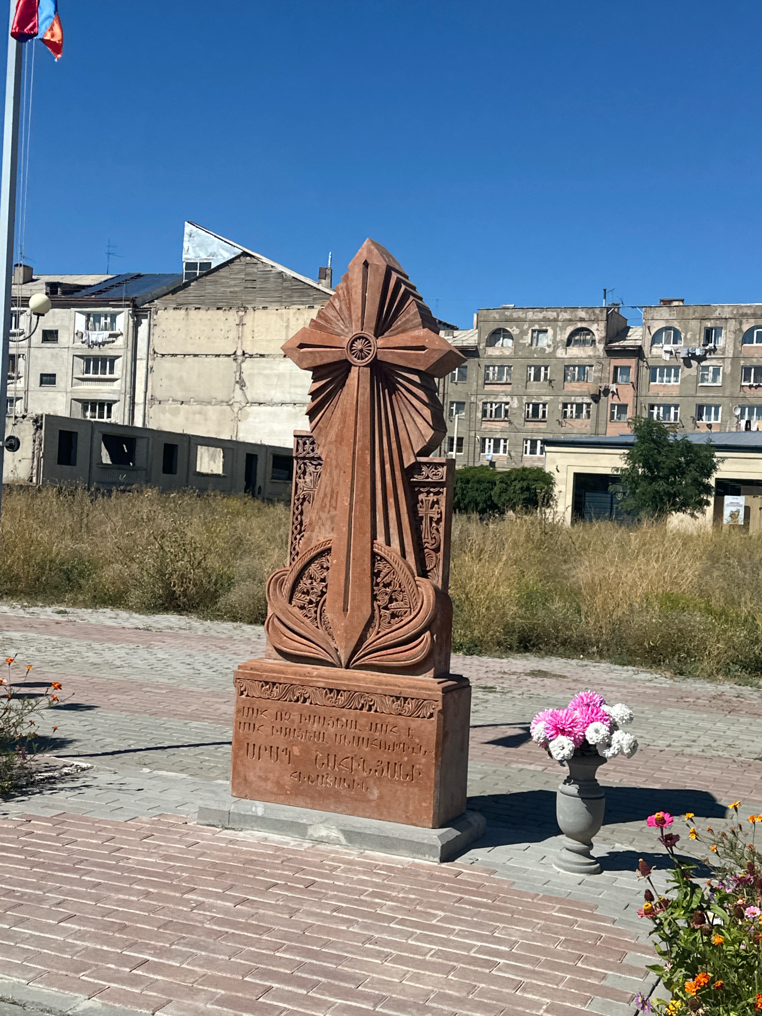This is a ground-level, eye-level, outdoor, full shot photograph of a monument in Gyumri, Armenia. The monument, made of reddish-brown stone, features a large cross with intricate carvings and an inscription in Armenian. In front of the monument, a grey urn holds a bouquet of pink and white flowers. To the right of the urn, a patch of wild flowers with pink, orange and yellow blossoms is visible. The monument and flowers are set against a backdrop of urban buildings, some appearing old and worn, and others more modern. The sky is clear and blue. The ground is paved with red and grey interlocking bricks. The overall scene suggests a public space, possibly a park or memorial site.