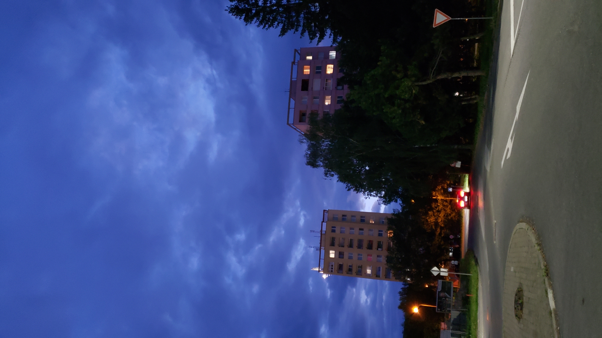 The image shows a twilight scene in the city of Prešov, Slovakia, featuring two apartment buildings, trees, a road, and a car. The sky is a deep blue with dramatic clouds. The apartment buildings are tall, with illuminated windows, suggesting it's evening. The building on the left is a light yellow color, and the building on the right appears to be pink. A dark green canopy of trees separates the road from the apartment buildings. The road is paved and appears to be an intersection or roundabout, with white lane markings and a give way sign visible. A red car with its brake lights illuminated is driving on the road. Streetlights are also visible, casting a warm glow.