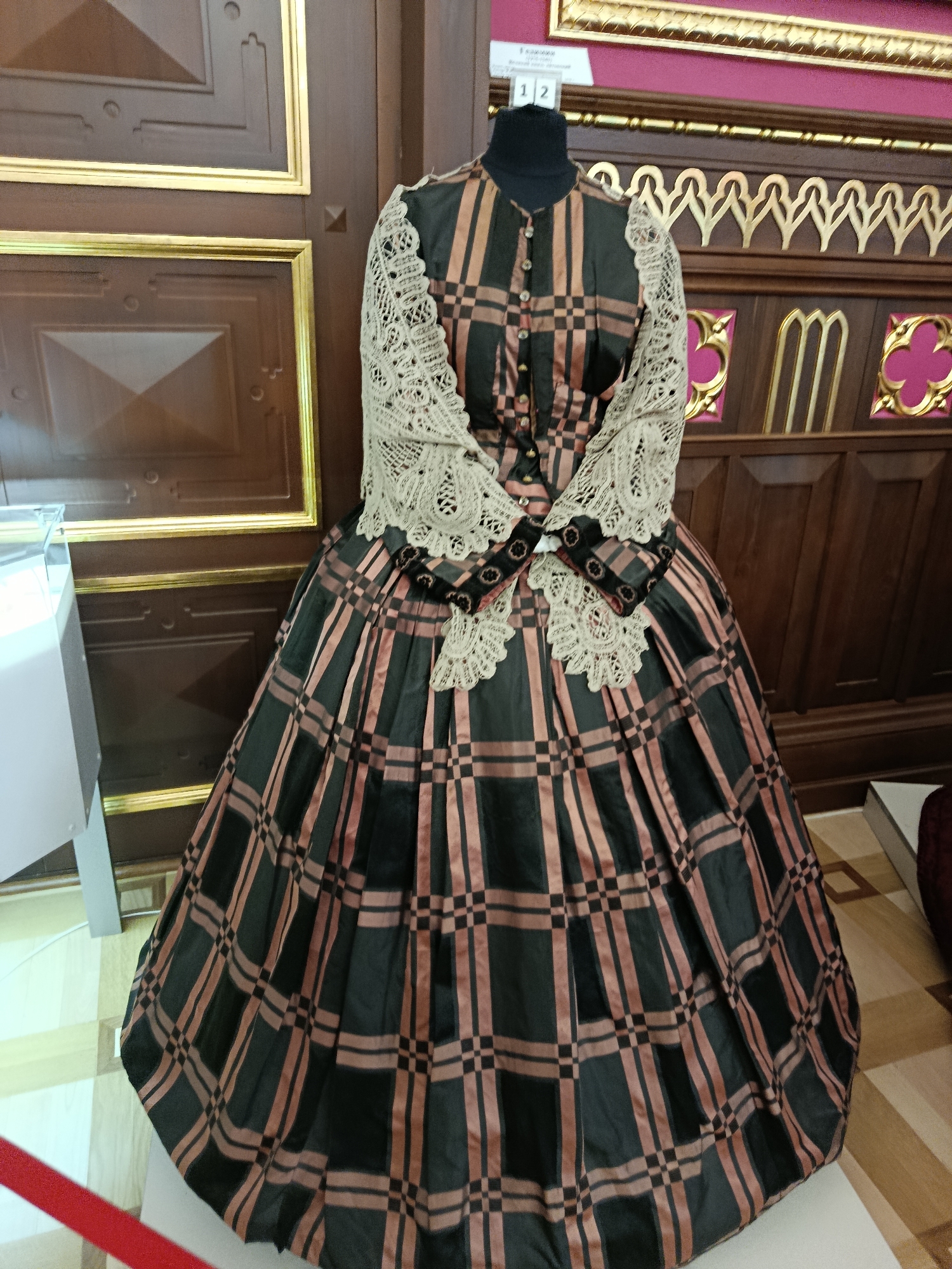 The image shows a mannequin dressed in an antique gown with a plaid pattern of dark green and tan. The dress has a fitted bodice with buttons down the front and a wide, flowing skirt. The bodice is adorned with lace trim around the neckline and sleeves. The background features ornate wooden paneling and decorative elements typical of a museum setting. This particular display is located within the Kosava Palace, a historical landmark in Kosava, Belarus.