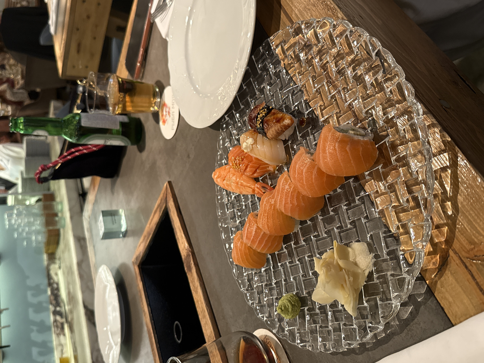 A close-up, overhead shot shows a table at a sushi restaurant in Moscow, Russia. On the table are several pieces of sushi, including salmon rolls and nigiri, arranged on a clear glass plate. To the left of the plate is a small mound of wasabi and some pickled ginger. Behind the sushi plate, a white plate sits empty, and a glass of amber liquid, likely beer, is next to a green bottle of beer. A dark cloth bag with a red drawstring is also visible. In the background, a bar area with shelves of bottles and glasses can be seen. The table appears to be made of dark stone, with a wooden border framing a dark inset area. The lighting is dim, creating a warm and inviting atmosphere.