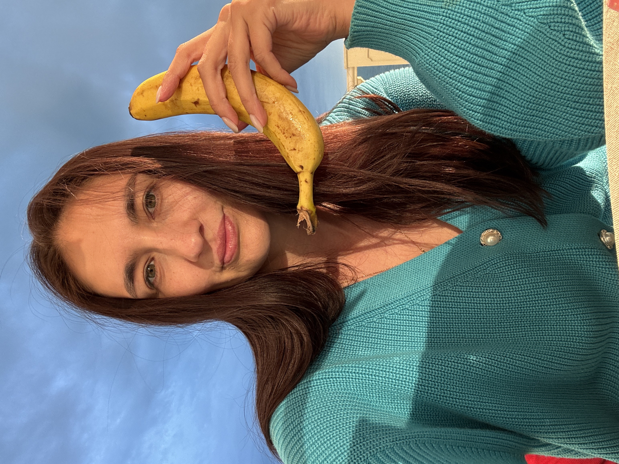 A person with auburn hair and green eyes is holding a ripe banana in their right hand, above their head. The person is wearing a turquoise knitted sweater or cardigan with pearl-like buttons. The background is a clear blue sky. The location is likely Sochi, Russia.