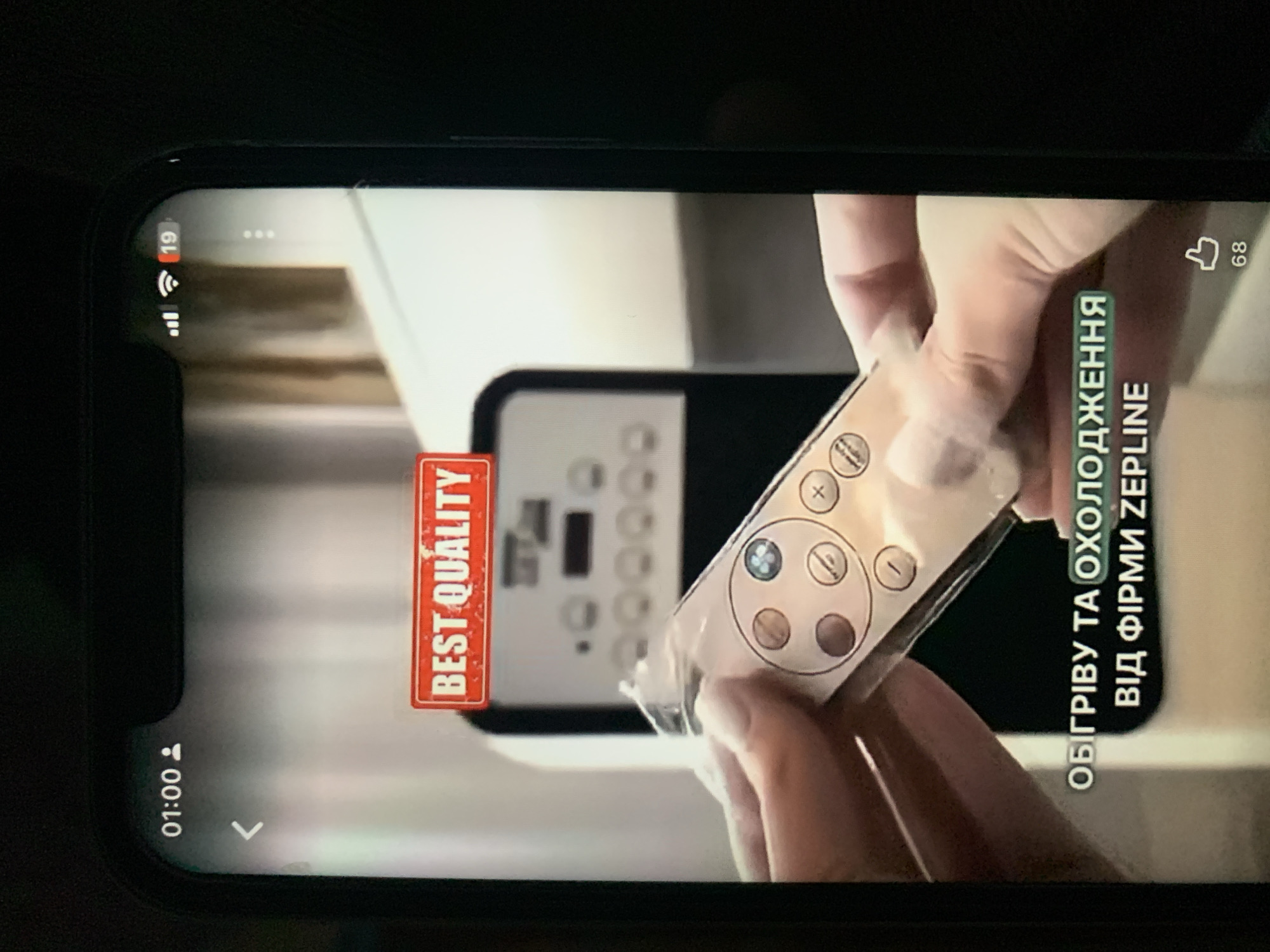 The image is a close-up shot of a smartphone screen displaying a video. The video shows a hand holding a clear plastic-wrapped remote control. In the background, a white appliance with buttons is visible. A red banner with white text reading "BEST QUALITY" is overlaid on the video. Below the appliance, text in Ukrainian reads "ОБІГРІВУ ТА ОХОЛОДЖЕННЯ ВІД ФІРМИ ZEPLINE". The top left corner of the phone screen displays "01:00" and a person icon, indicating the time and possibly a user profile. The top right corner shows signal strength, Wi-Fi icon, and a notification bubble with the number "19". At the bottom right, a thumbs-up icon with the number "68" is visible, likely representing likes. The scene is not identifiable as being in Zhmerynka, Ukraine, as no geographical markers are present in the image.