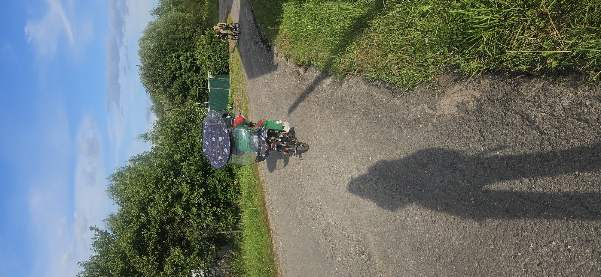 In Vitsebsk, Belarus, on a rural asphalt road bordered by lush green grass and trees, a person rides a motor scooter equipped with a large, transparent canopy. The canopy, adorned with a dark blue fabric patterned with white stars, extends over the rider and the back of the scooter, providing protection from the elements. The rider is wearing a red top with green stripes down the side of their pants. In the distance, two individuals on bicycles are also visible on the same road. The sky above is a clear blue with scattered white clouds, and a long shadow of the person taking the photo stretches across the road in the foreground. A green metal fence runs along the edge of the property behind the scooter.
