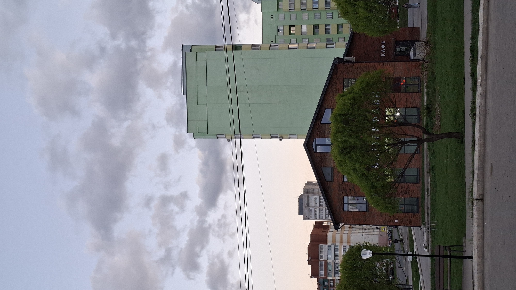 The image presents an urban scene in Chaikovsky, Russia. The foreground features a red brick building with a pitched roof, identified by signage as a "Cafe" in Russian. A mature willow tree stands in front of the building. A green apartment building rises behind the café. In the background is a sky filled with clouds. A streetlight and pedestrian bench suggest a park-like setting. Power lines are present.