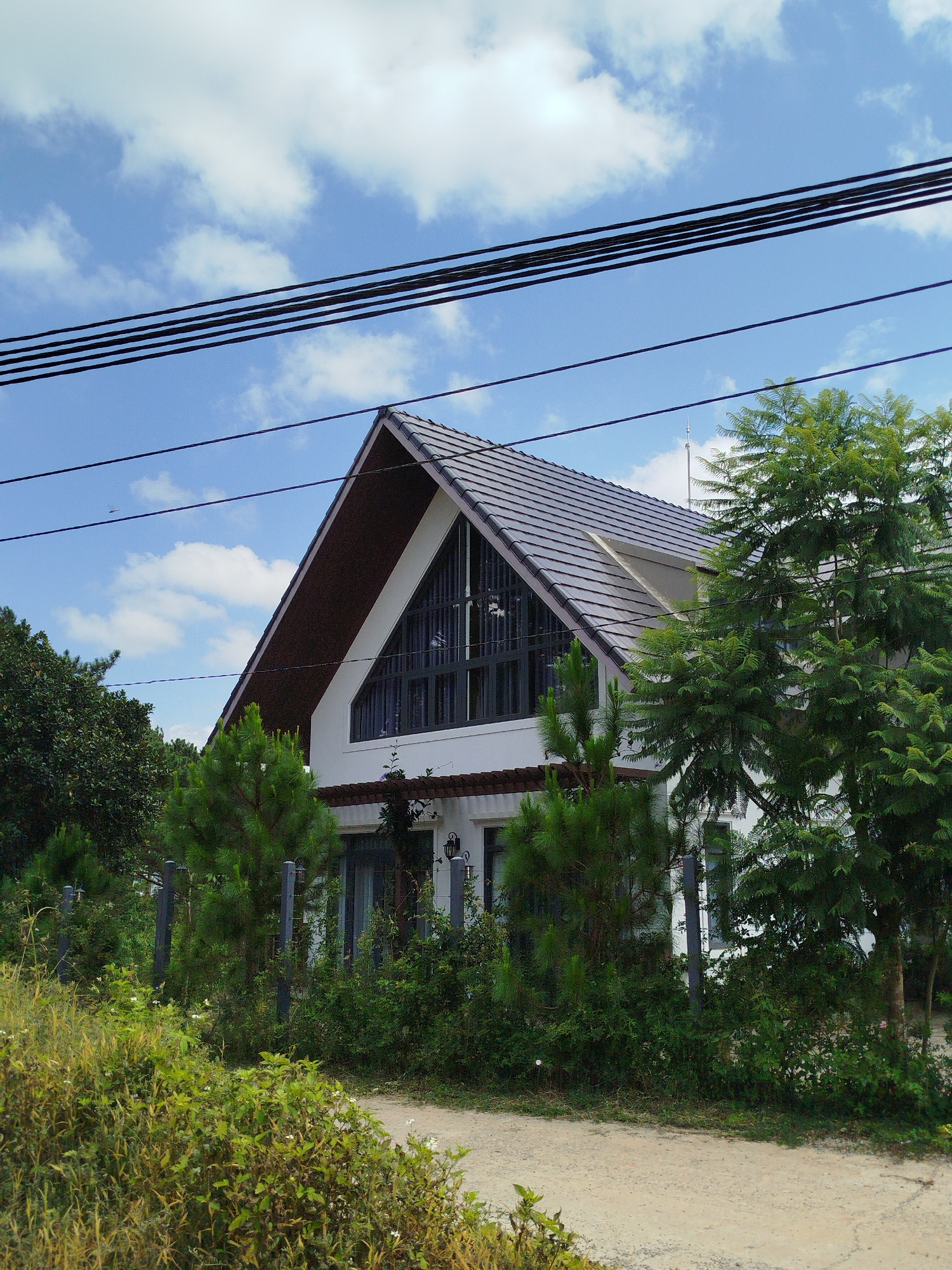 The image depicts a modern white house with a dark gray tiled roof, featuring a prominent triangular-shaped window on its upper facade. The house is surrounded by lush green trees and vegetation, with a dirt path leading towards it. Several power lines are visible against a backdrop of a bright blue sky dotted with white clouds. The setting appears to be a quiet, possibly rural or suburban area within the city of Tuý Sơn, Vietnam, given the presence of dense greenery and overhead power lines. The scene is tranquil and shows a well-maintained property integrated with its natural surroundings.