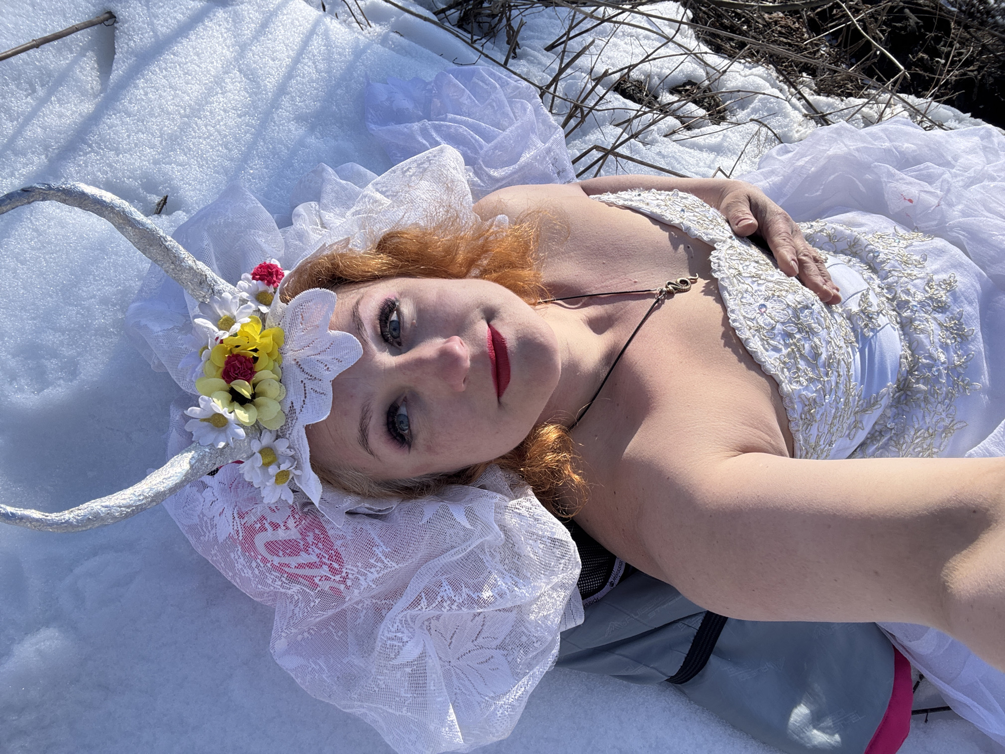 A woman is lying on her back in the snow in a sunny, outdoor environment. She is dressed in a white gown with gold embroidery, and a lace headpiece adorned with flowers and silver horns. Her face is tilted towards the camera, and she is wearing red lipstick. Her hair is reddish-blonde. Her right hand is resting on her chest. Part of a grey and black bag is visible beneath her. In the background, there is bare earth with some twigs and branches. It is daytime, and given the prompt, it is implied that this scene is located in Chaikovsky, Russia.