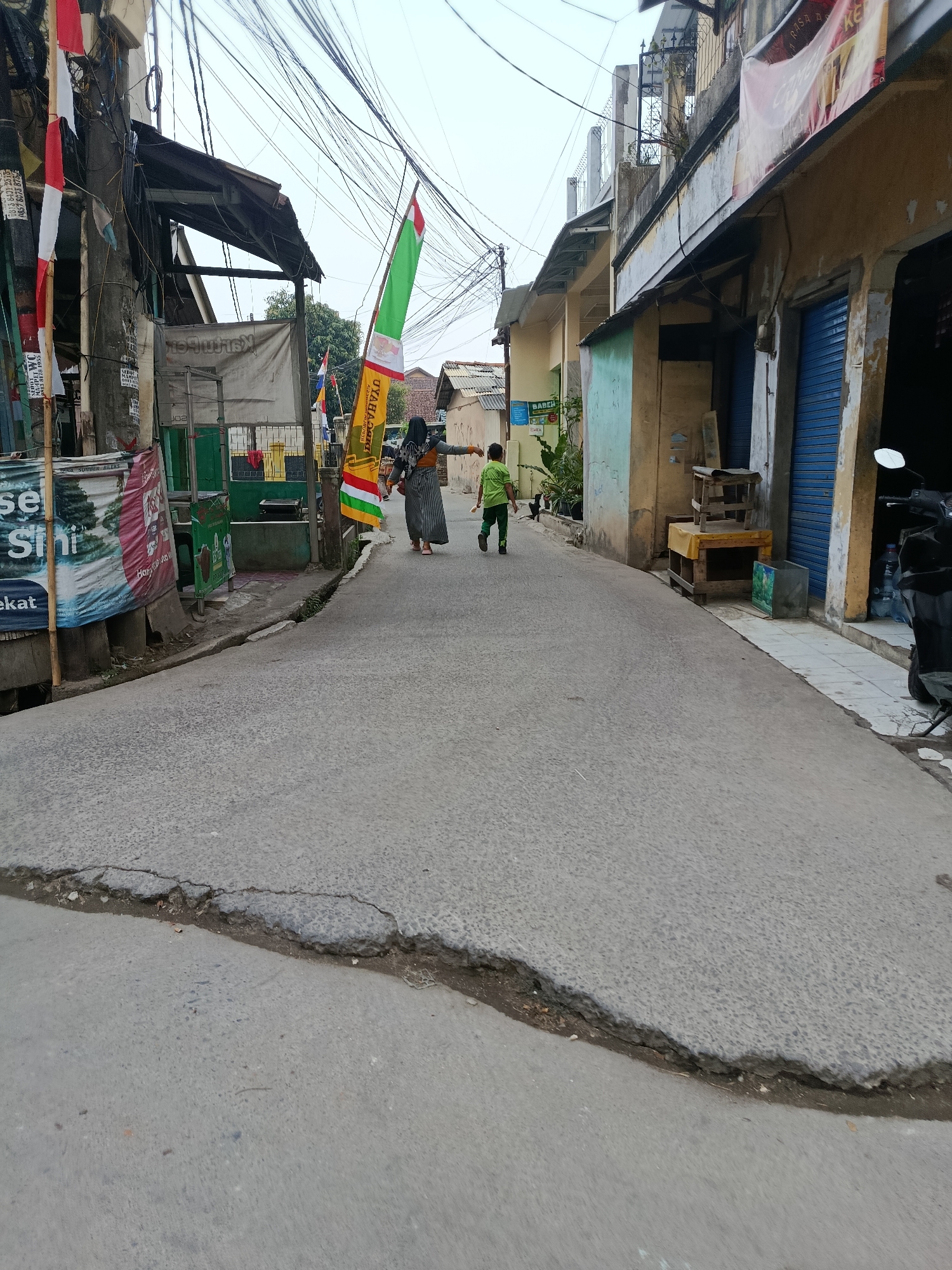 The image shows a narrow street in Balaraja, Indonesia, lined with buildings and overhead electrical wires. The street surface is asphalt and appears cracked in places. Two individuals are walking away from the camera. One is an adult wearing a long patterned dress and a headscarf. The other is a child in a green shirt and pants. Various flags, including Indonesian flags, are displayed along the street. One building on the right has blue metal shutters. A motorcycle is parked on the right side of the street. The sky is overcast.