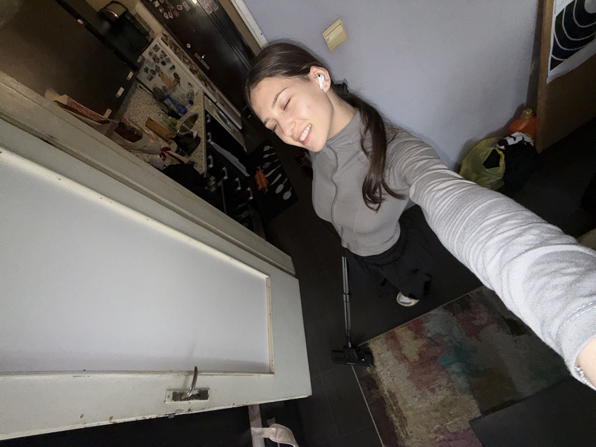 A smiling young woman with dark hair, wearing a gray long-sleeved shirt and black pants, is taking a selfie. She has an AirPod in her left ear. The location appears to be indoors, potentially a kitchen or living area. A white door or cabinet is visible in the foreground, and a vacuum cleaner is on the floor. A rug is partially visible. The location in the city of Sofia, Bulgaria, cannot be ascertained from the image.