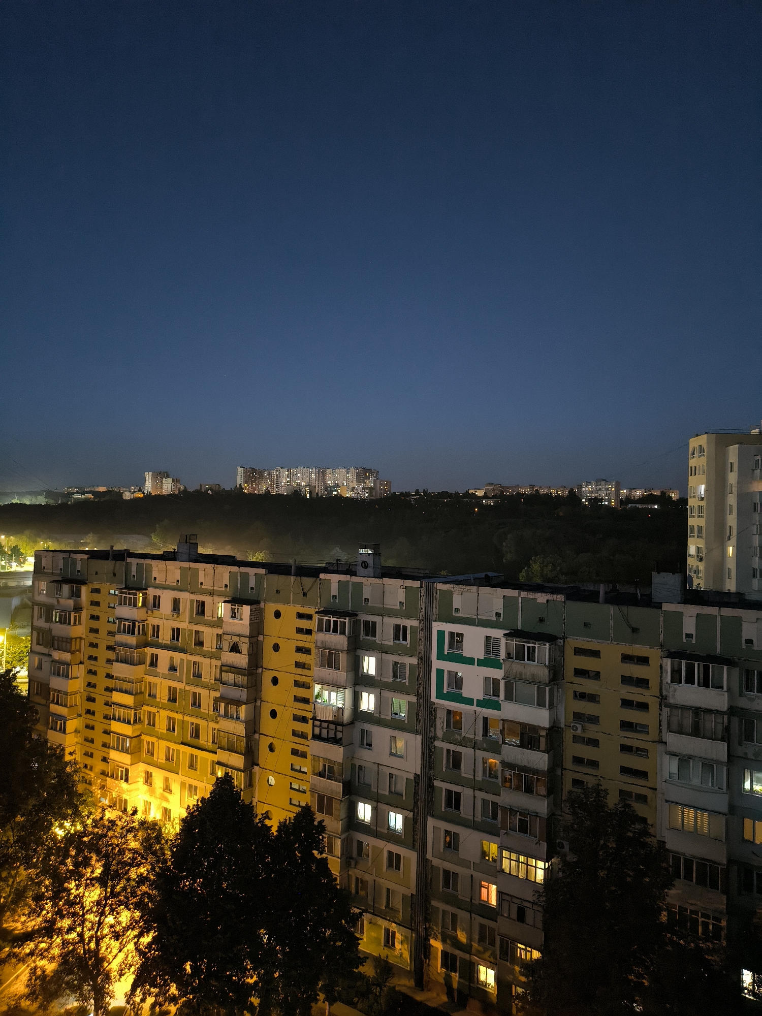 This is a night-time, slightly elevated, wide-angle photograph of a residential area in Chișinău, Moldova. The scene is dominated by several multi-story apartment buildings. In the foreground, a large apartment building with a yellow and light green facade is prominent. It features numerous balconies and windows, many of which are illuminated, suggesting occupancy. Trees, some with illuminated foliage from streetlights, are positioned in front of this building.
Behind the foreground building, a dense line of trees forms a dark, undulating band across the mid-ground, obscuring the lower portions of distant structures. Beyond the trees, in the background, several more apartment buildings are visible, illuminated with countless lights, indicating a larger urban settlement. These buildings are of varying heights and architectural styles, suggesting a mix of Soviet-era and possibly newer construction.
The sky above is a deep, dark blue, characteristic of twilight or early night, with a few faint stars visible. The overall atmosphere is quiet and residential, with the artificial lights of the apartments and streetlights creating a warm glow against the dark sky and landscape. The perspective suggests the photograph was taken from another building or a higher vantage point, looking out over the cityscape.