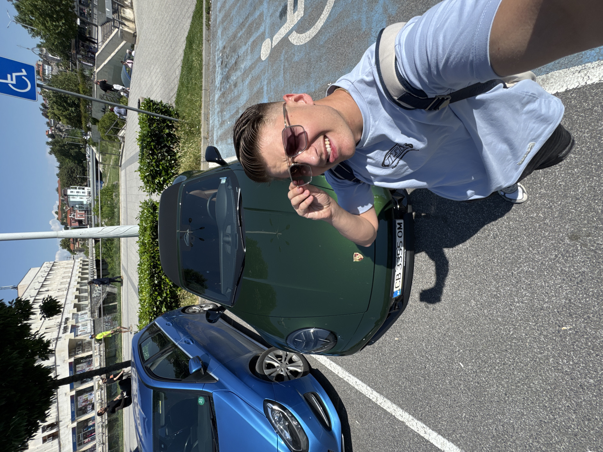 A man is taking a selfie while standing next to two cars parked in a parking lot. The man is wearing a light blue t-shirt, sunglasses, and a shoulder bag. He is smiling and holding his sunglasses with one hand. One car is dark green and appears to be a Porsche convertible, with the license plate "CB 3355 OM." The second car is blue and partially visible to the left of the green car. The parking spot features a handicap parking sign, and the parking space is marked with the corresponding symbol. In the background, there are buildings and trees, indicating an urban environment. The photo was taken during the day, with bright sunlight. It is likely that the photo was taken in the city of Plovdiv, Bulgaria, as the license plate indicates that it is registered in the Plovdiv district.