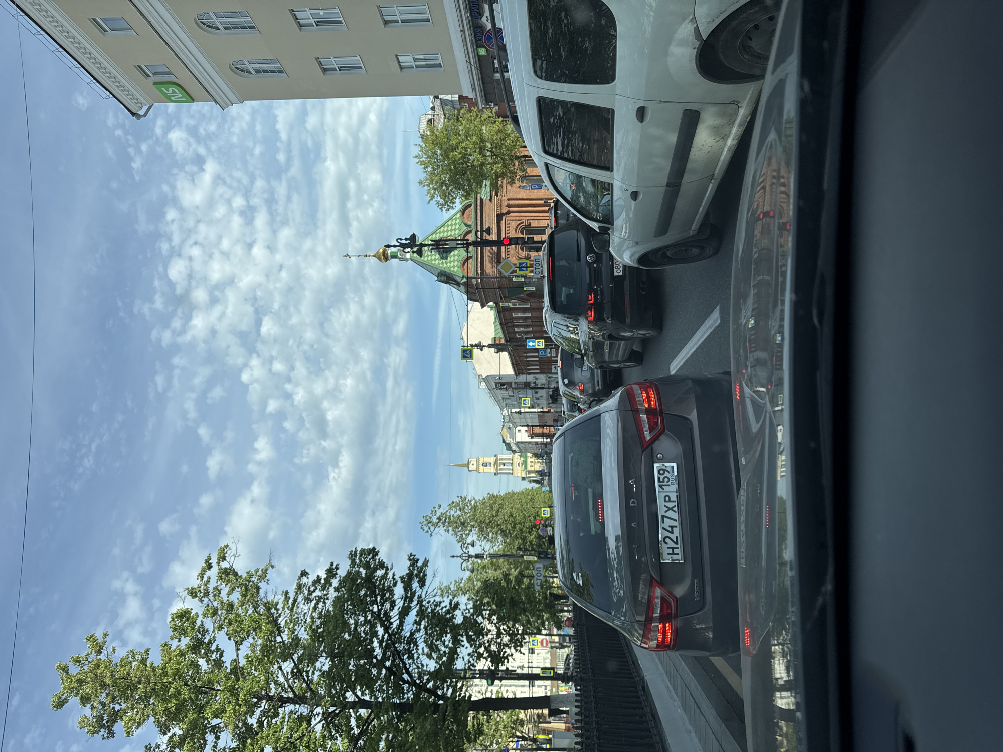 The image captures a street scene in Perm, Russia, characterized by a traffic jam. Prominent in the foreground is the rear of a dark-colored Lada Vesta car with the license plate H247XP 159 RUS. The image is taken from inside a vehicle, likely another car, evidenced by the frame of the window on the right side. The presence of other vehicles indicates a congested road, including a white van in front.
In the background, a distinct architectural feature is visible: a brick church with a green roof and a golden dome. Further down the street, a slender, light-colored tower can be seen.
The sky is partially visible, showing a mix of blue sky and scattered white clouds. Green trees line the side of the road, adding to the urban environment. The image gives a sense of a typical day in the city, with everyday traffic and elements of local architecture.