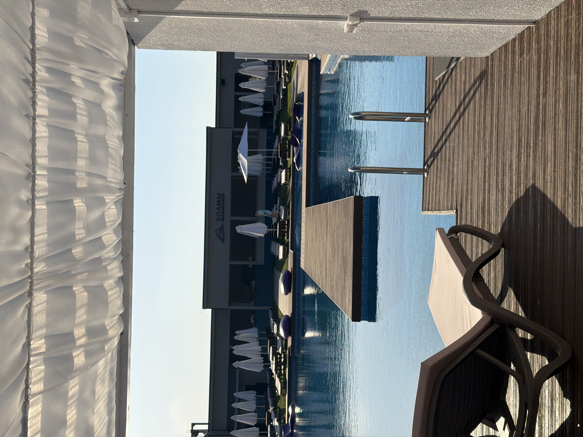 The image, when oriented correctly, presents a tranquil outdoor swimming pool area at a resort or hotel, identified by the visible text "ХОЛМЫ" (KHOLMY, meaning "Hills") on a building in the background. Given the location context, this is likely in Дрокино, Russia.
The scene is illuminated by warm, soft light, indicative of either early morning or late afternoon, as evidenced by the long, distinct shadows cast by structures like the pool railings onto the wooden deck. On the left side of the frame, a white, semi-transparent fabric, possibly a curtain from a cabana or shaded lounge, drapes downwards, catching the sunlight and forming gentle undulations. To the right, a dark, intricately designed lounge chair with a light-colored cushion rests on a textured wooden deck that lines a portion of the large, inviting blue swimming pool.
Metal railings, likely part of a pool ladder, extend into the clear, blue water. Across the pool, the main resort building, featuring a dark, modern facade, forms the backdrop. In front of this building, several rows of white beach umbrellas and corresponding lounge chairs are neatly arranged along the poolside, suggesting areas for relaxation and sunbathing. Purple, spherical decorative objects are also interspersed along the pool's edge in the background. A small, dark wooden platform extends into the water from the far side of the pool, adding a design element. The cloudless sky is reflected beautifully in the calm pool water, contributing to the serene and leisurely atmosphere of the setting.