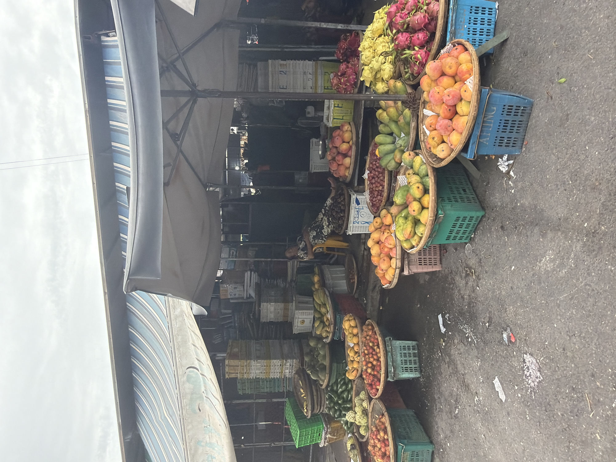 A bustling fruit stand in Nha Trang Ward, Vietnam, is overflowing with a colorful assortment of fruits displayed in woven baskets and plastic crates. The stand is shaded by an awning with a blue and white striped pattern. A variety of fruits including dragon fruit, mangoes, bananas, and custard apples are on display. A vendor sits on a yellow stool behind the fruit stall, with stacks of empty white and yellow crates visible in the background. The ground is concrete with some debris and dirt scattered around.