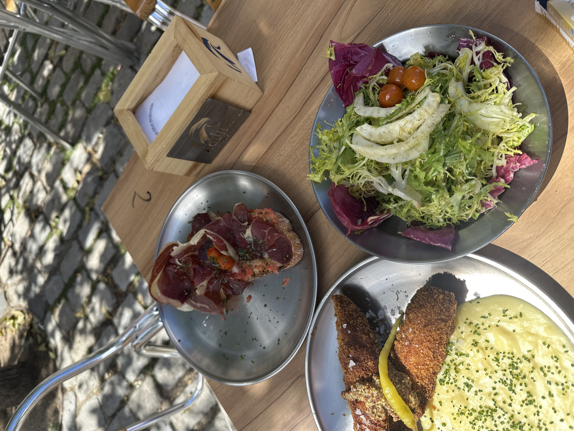 The image shows a table setting with three metal plates of food. One plate contains a salad with green lettuce, red lettuce, fennel, and cherry tomatoes. Another plate holds a slice of bread topped with prosciutto and tomatoes. The third plate contains fried food accompanied by mashed potatoes sprinkled with chives and topped with pepper. A wooden napkin holder sits on the table, and there is a metal chair next to the table. The number "2" is visible on the table. The setting is consistent with outdoor dining in Madrid, Spain.