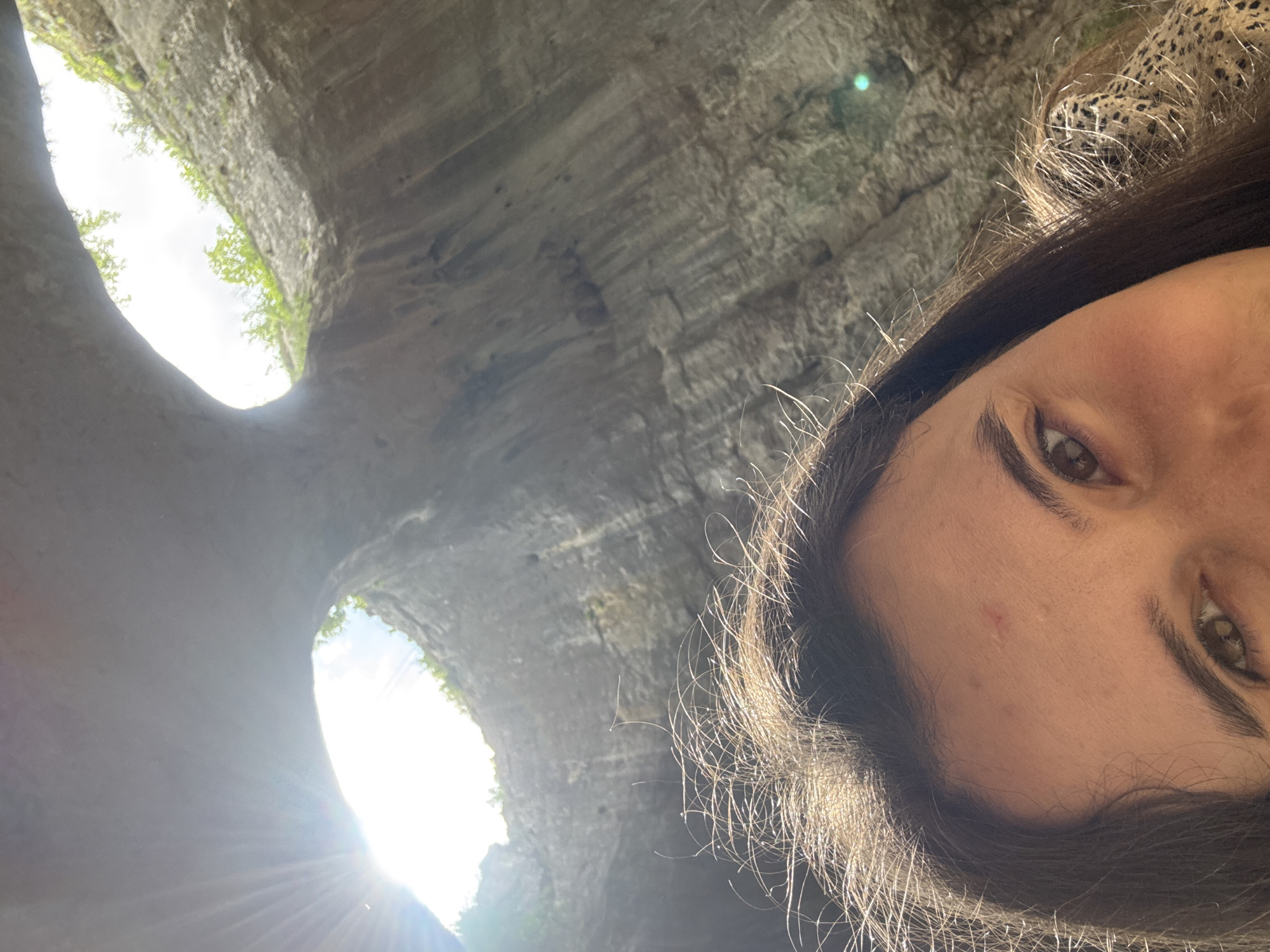 A close-up, slightly angled selfie-style photograph depicts a woman's face in the foreground, positioned towards the right side of the frame. Her eyes, hair, and a portion of her patterned clothing are visible.
Behind the woman, occupying the left side and background of the image, is a geological feature. It appears to be a cave or rock formation with a natural opening through which daylight streams. The light creates bright flares and illuminates the interior of the structure. The location appears to be the rock formation "Prohodna Cave", located in Karlukovo, Bulgaria.