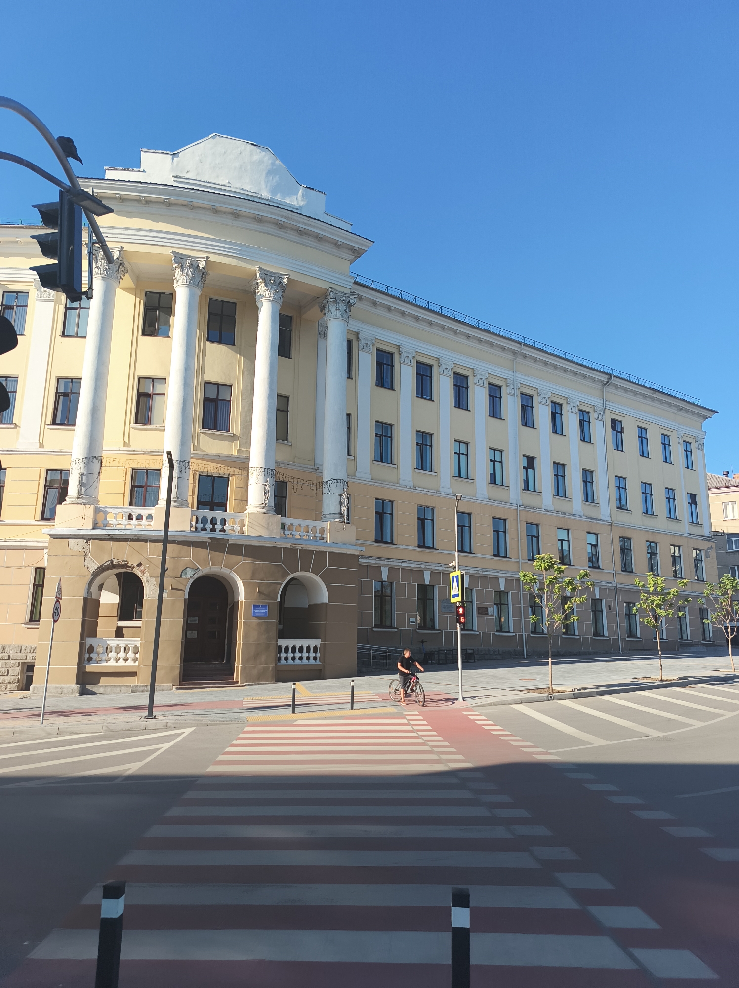 This is a daylight exterior shot of a large, light yellow building with classical architectural elements, such as columns and a pediment, located in Ternopil, Ukraine. The building has multiple stories and numerous windows. In the foreground, a pedestrian crossing with red and white stripes is visible, along with a road and sidewalk. A person on a bicycle is crossing the street on the pedestrian path, and a traffic light is visible. Trees are planted along the sidewalk in front of the building. The sky is clear and blue.