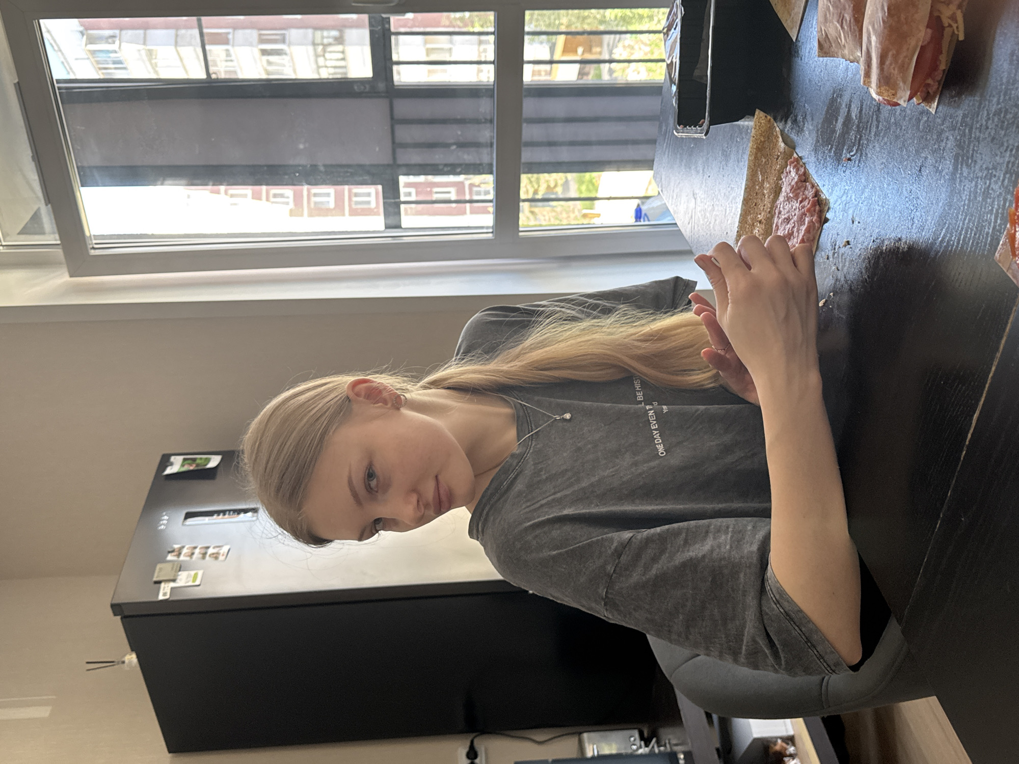A young woman with blonde hair is seated at a black table indoors, possibly in an apartment in Perm, Russia. The room is illuminated by natural light coming through a window. The window reveals a view of a multi-story building across the street. The woman is holding a wrap, likely with meat inside. On the table, there are other wraps. To her left, a black refrigerator is visible. The woman is wearing a gray t-shirt and a necklace.