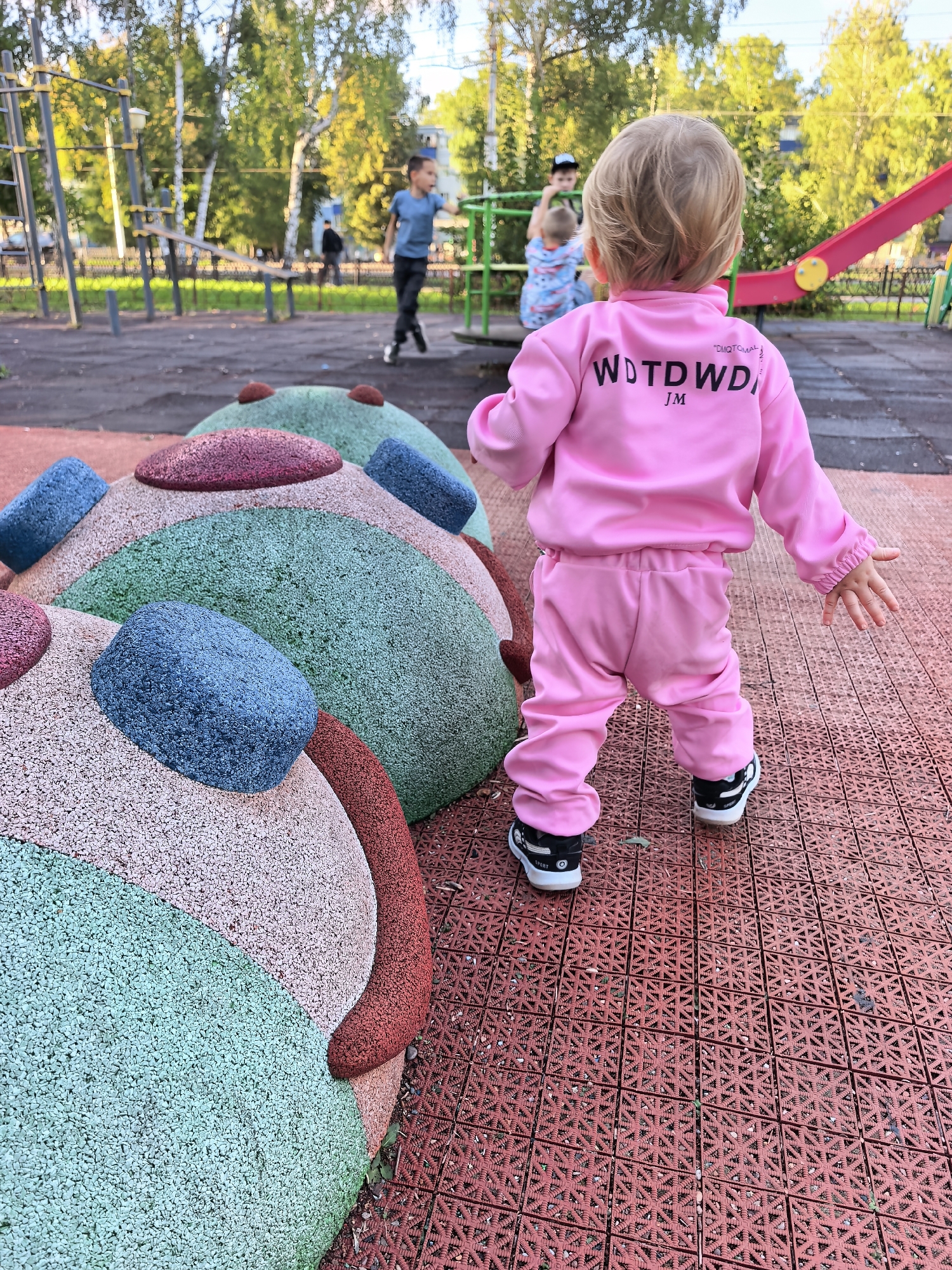 In Sterlitamak, Russia, a young child in a pink tracksuit stands on a red rubber playground surface. The child faces away from the camera, looking towards a colorful playground structure with a red slide visible in the background. The playground features soft, rounded mounds covered in multi-colored gravel. Other children can be seen playing in the background, with trees and a building visible beyond the playground. The scene captures a moment of outdoor play on a sunny day.