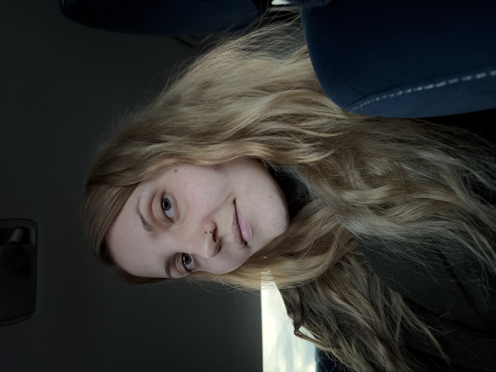 A young, light-skinned woman with long blonde hair, a nose ring, and light eyes looks at the camera with a slight smile. She is sitting in the front seat of a vehicle with dark blue upholstery. The interior of the vehicle is mostly dark except for a bright rectangle visible through the window that could be the sky. The location in Chaikovsky, Russia is unknown.